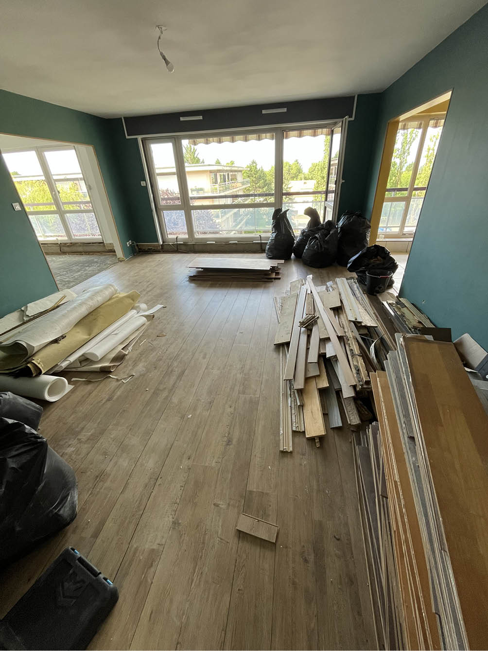 Room with large windows under renovation, with wood planks, rolled-up carpet, and black trash bags on wooden floor.