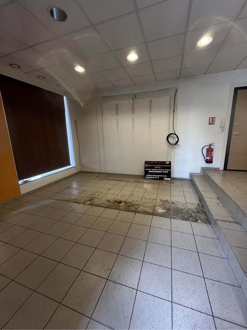Empty tiled room with a dirty patch on the floor, a closed dark window blind, stairs on the right, and a fire extinguisher mounted on the wall.