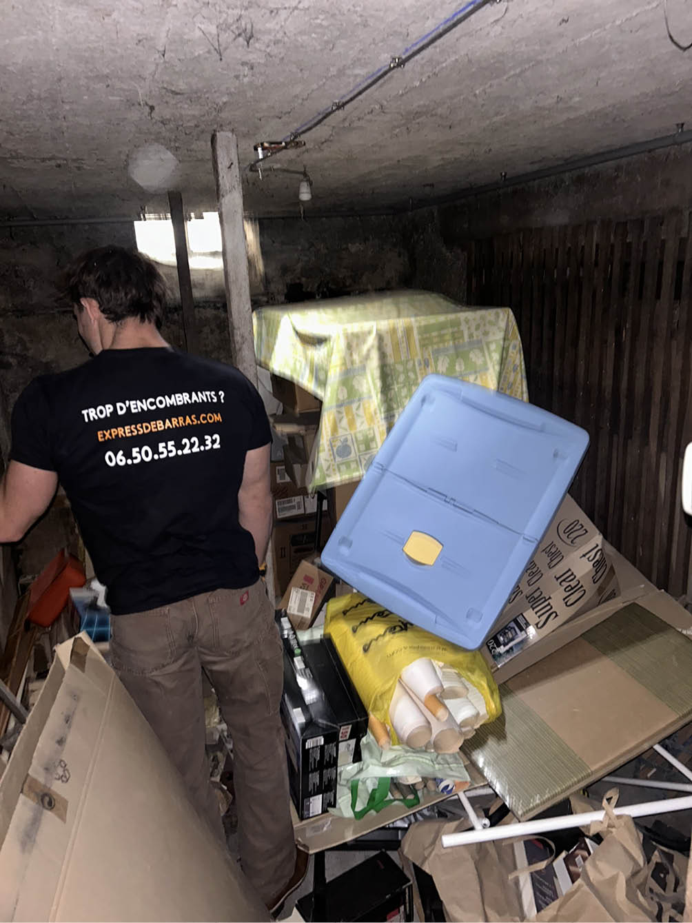 Man in brown pants and black shirt standing in a cluttered basement with boxes, bags, and a blue plastic container.