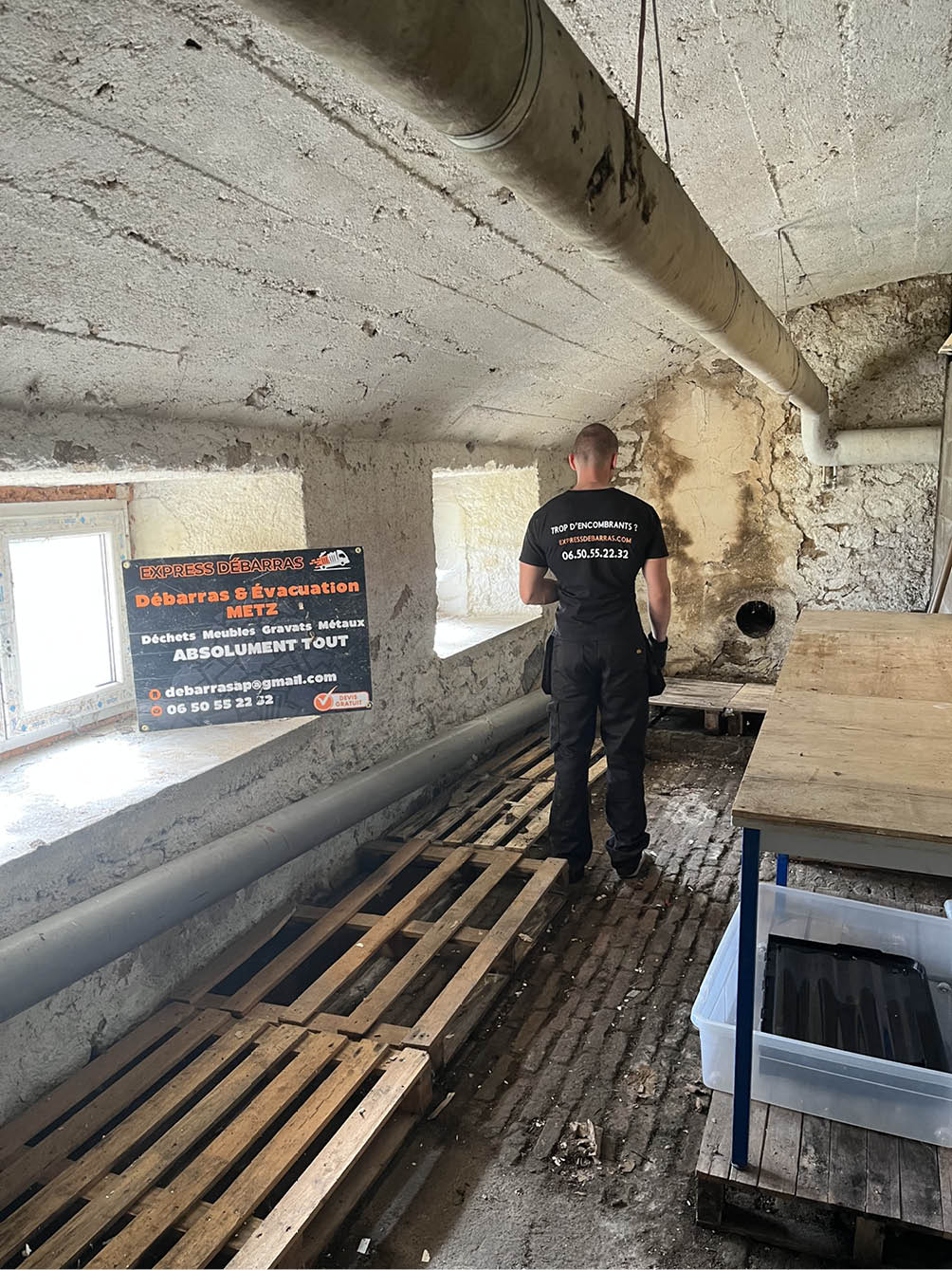 Person wearing black work clothes standing in a rustic attic with exposed brick floor and pipes, near a sign for debris removal services.