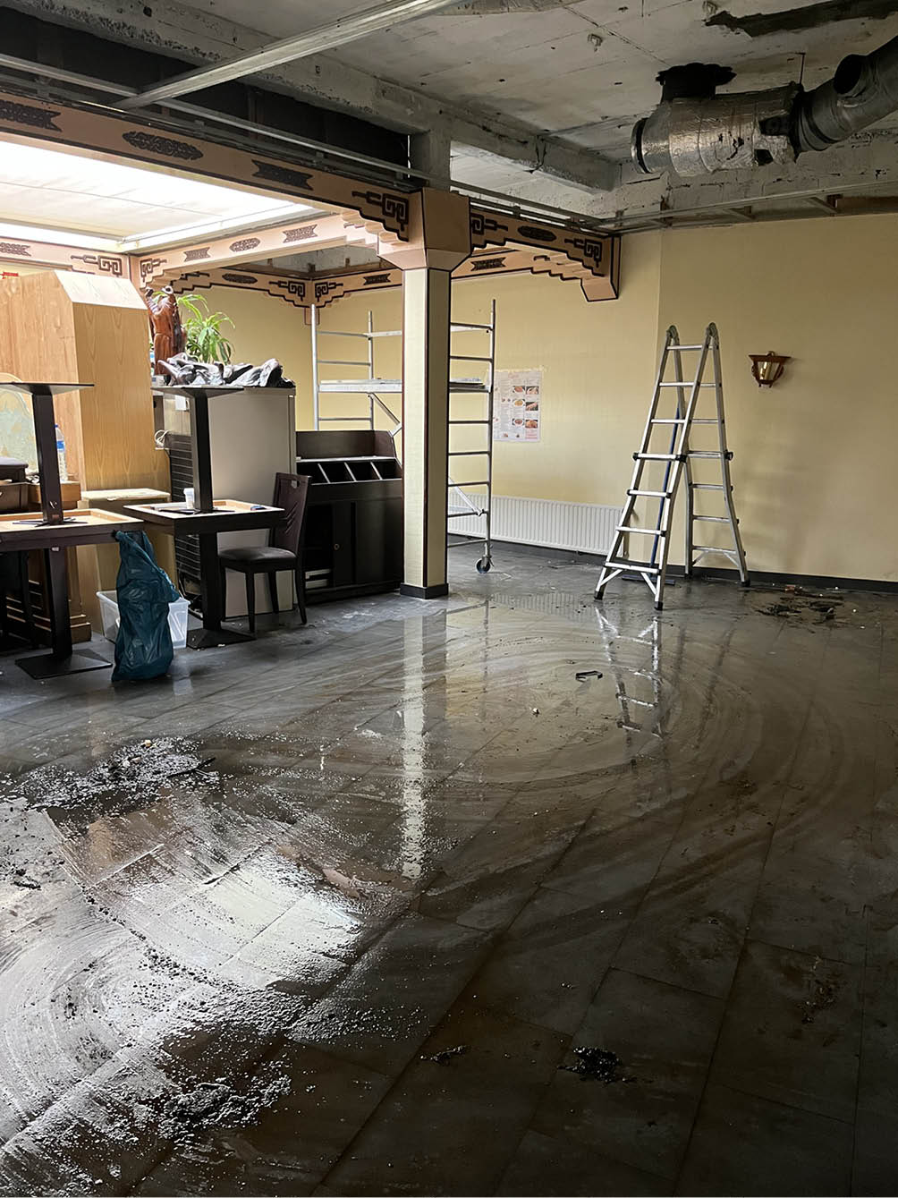 Indoor room with wet tiled floor, two ladders, scattered furniture, and exposed ceiling pipes.
