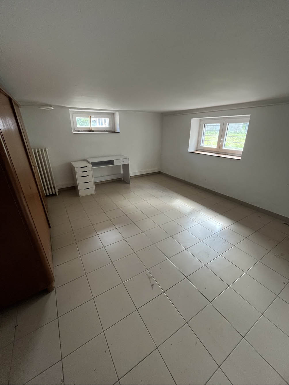 Empty room with white tiled floor, two small windows, a white desk with drawers, and a wooden wardrobe.