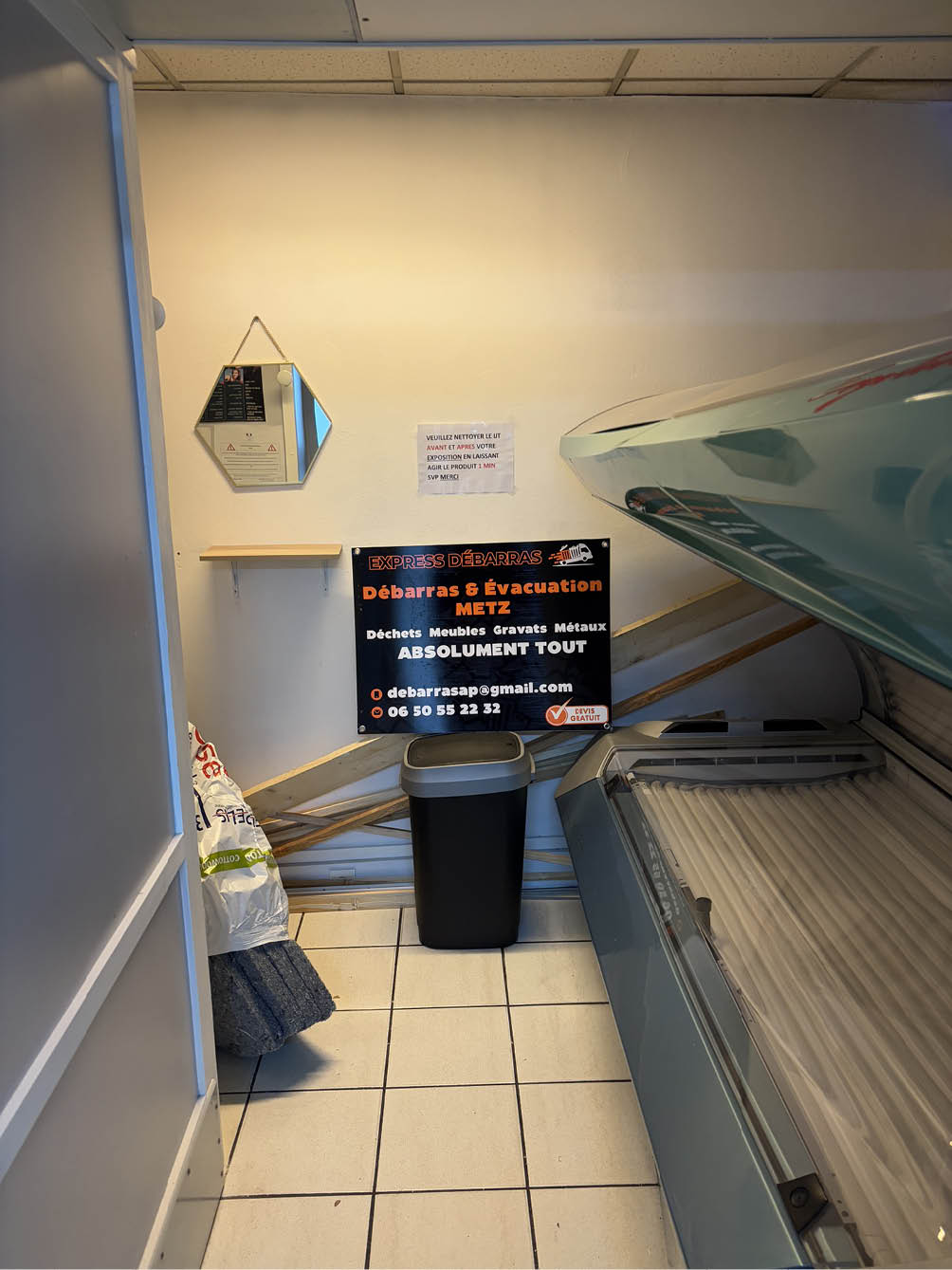 Small room with a tanning bed on the right, a black trash bin against the wall, a small shelf, a hexagonal mirror, and a sign advertising a clearing and evacuation service in Metz.