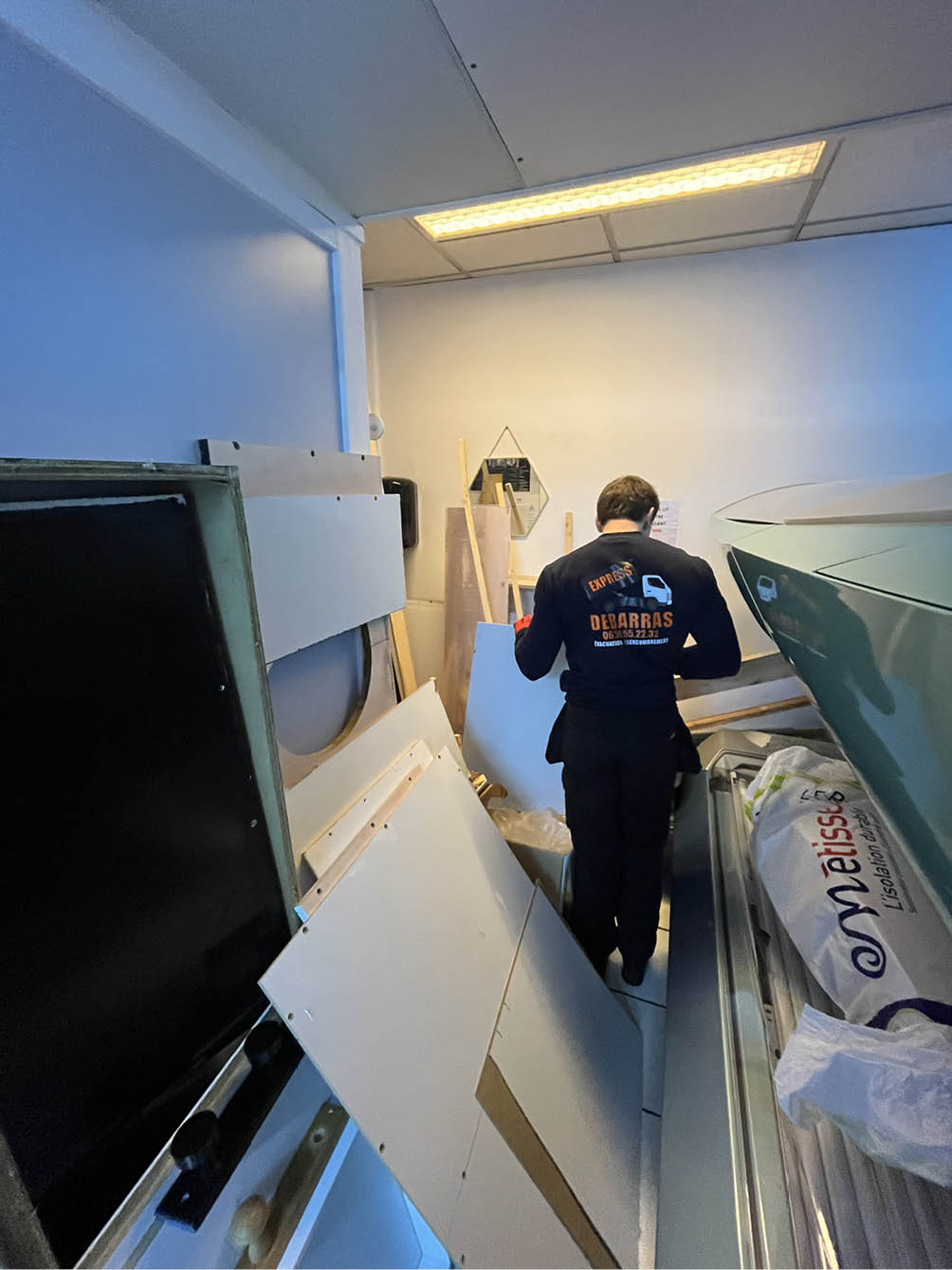 Person in dark clothing standing in a cluttered room filled with white panels and construction materials.