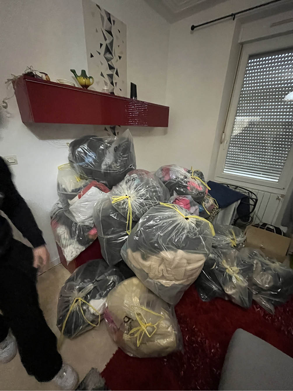 Room corner with multiple large transparent plastic bags tied with yellow strings, filled with clothes, placed on the floor near a red shelf and a window with closed blinds.