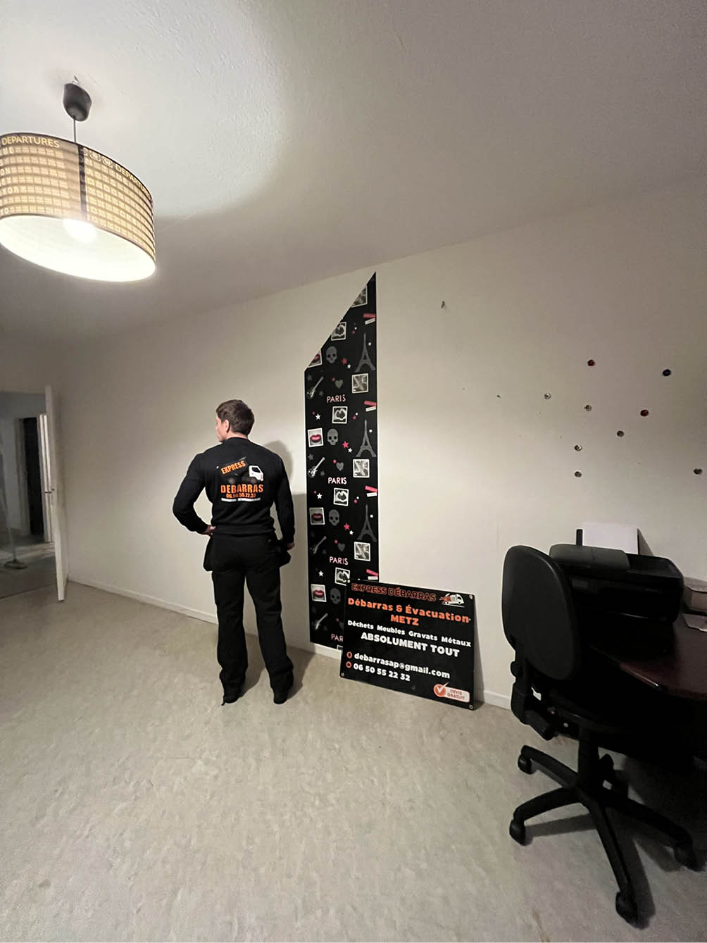 Man in black uniform standing in an empty room with a sign for Débarras & Évacuation Metz and a partially removed Paris-themed wall decal.