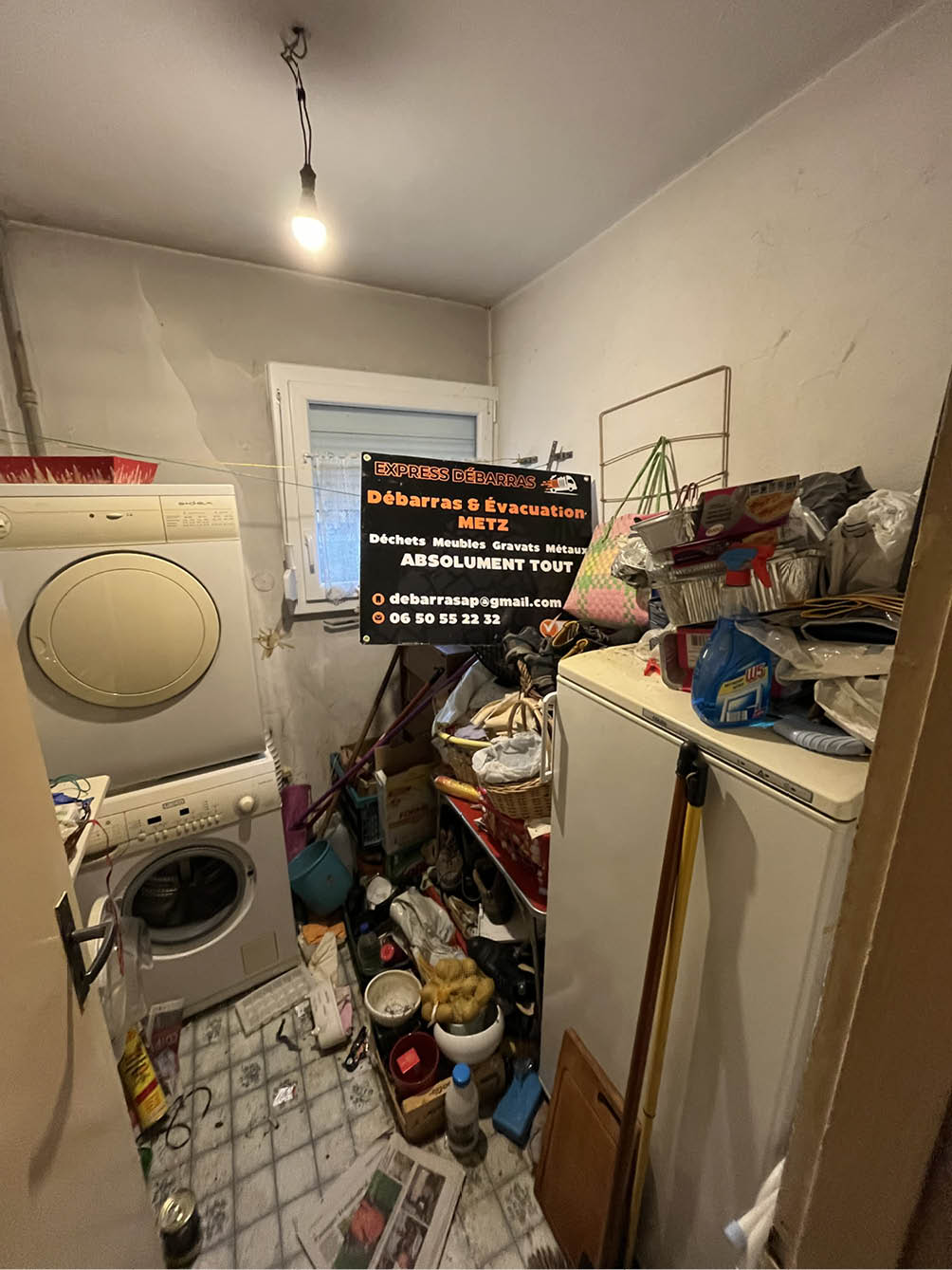 Cluttered laundry room with stacked washer and dryer, shelves filled with various items, cleaning supplies, and a sign for a junk removal service.