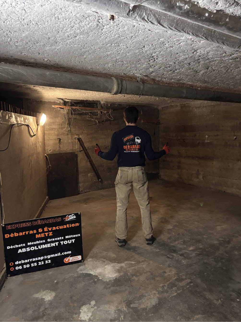 Man standing with thumbs up in an empty, dimly lit concrete basement room with an Express Débarras sign advertising clearance services.