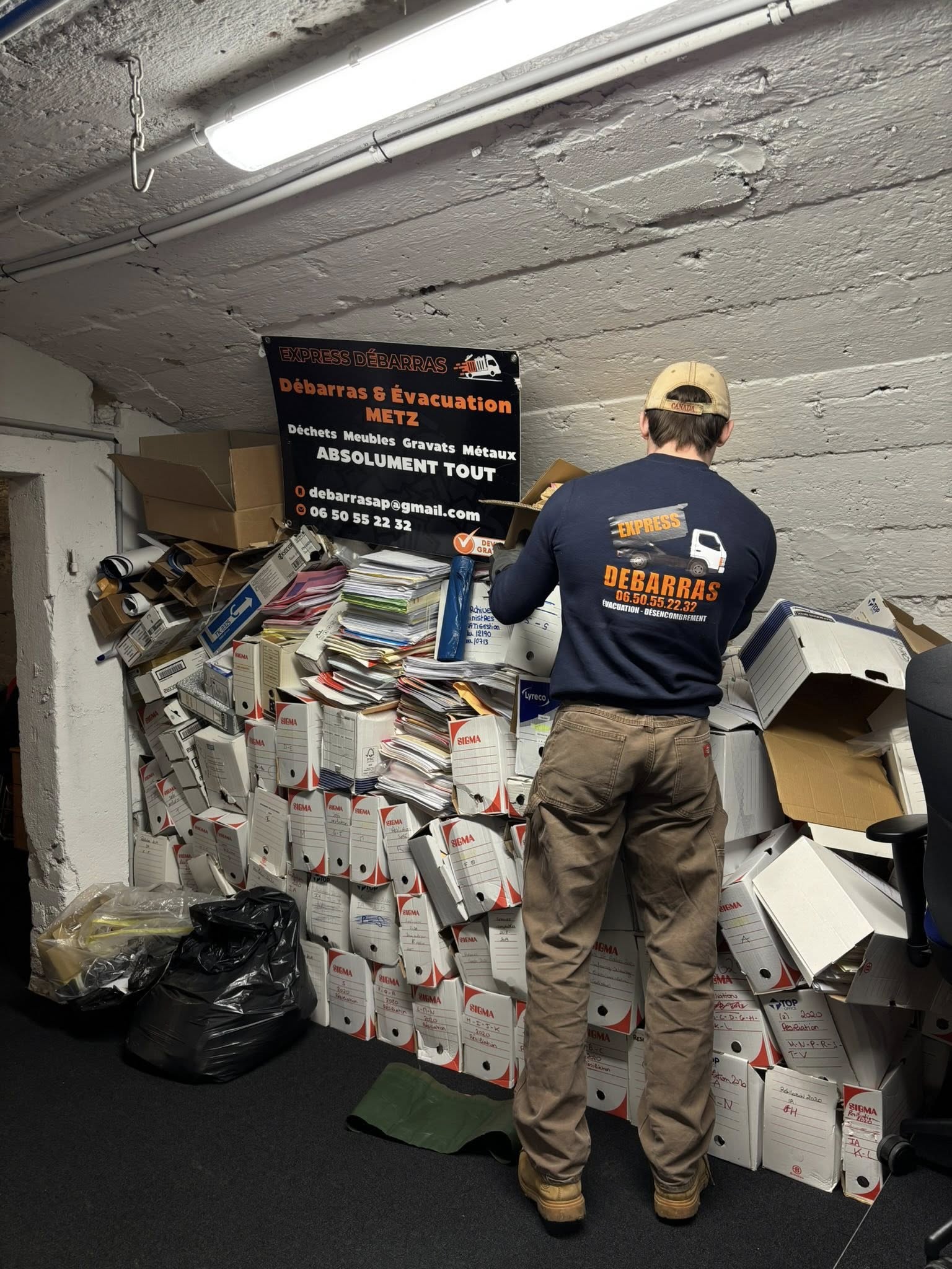Man wearing an Express Débarras uniform organizing a large pile of stacked document boxes and papers in a basement room.