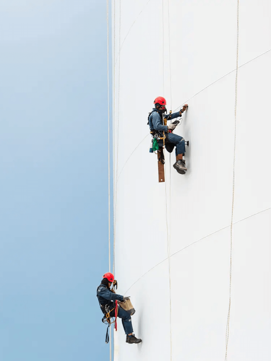 Zwei Arbeiter mit roten Helmen klettern an einem großen, weißen Tank entlang, gesichert durch Seile.