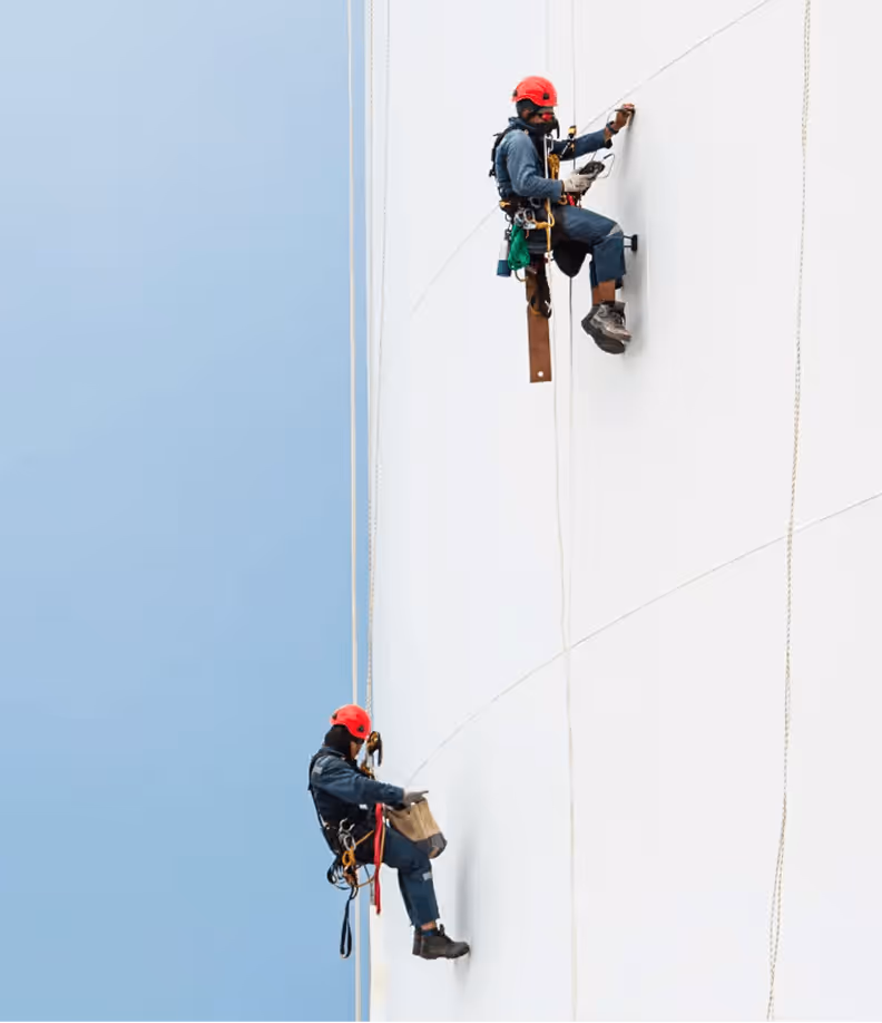 Zwei Arbeiter in roten Helmen klettern an einem weißen Industriebau und führen Wartungsarbeiten mit Seilen aus.