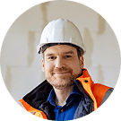 Bauarbeiter mit Helm und orangener Sicherheitsjacke lächelt in einer Baustellenumgebung.