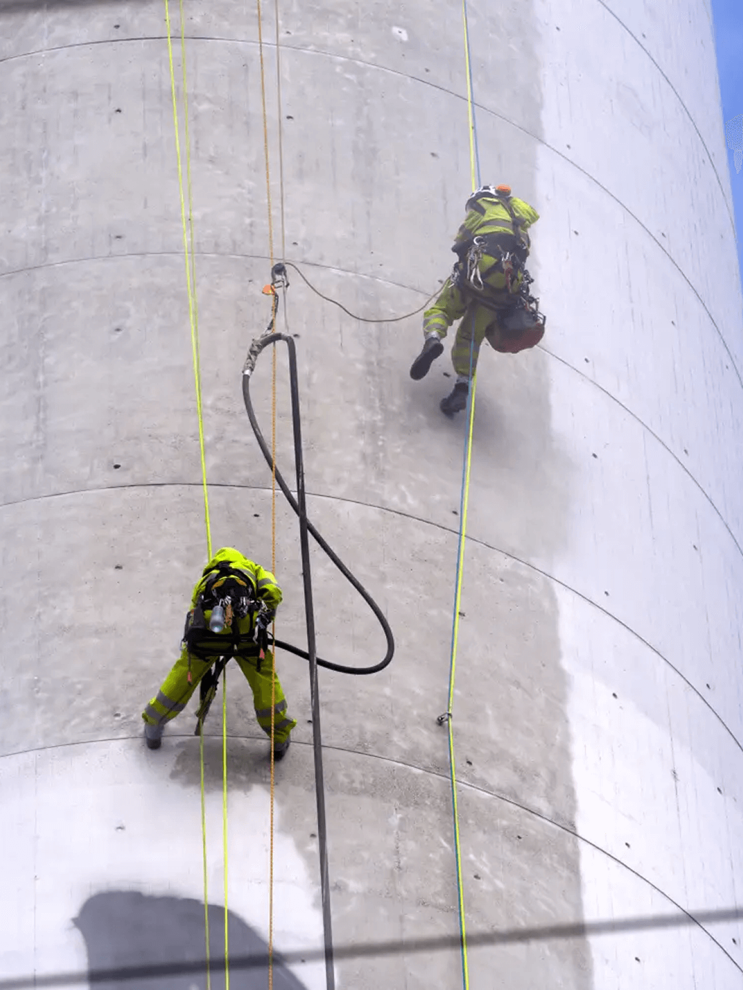 Industriekletterer in gelber Schutzkleidung arbeiten an einem großen Betonsilo mit Seilen und Spezialausrüstung.