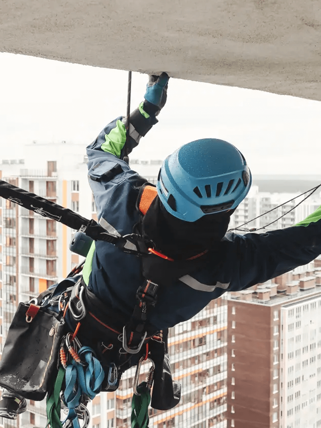 Arbeiter in Schutzkleidung und Helm hängt an Seil an Gebäudefassade, Hochhäuser im Hintergrund.
