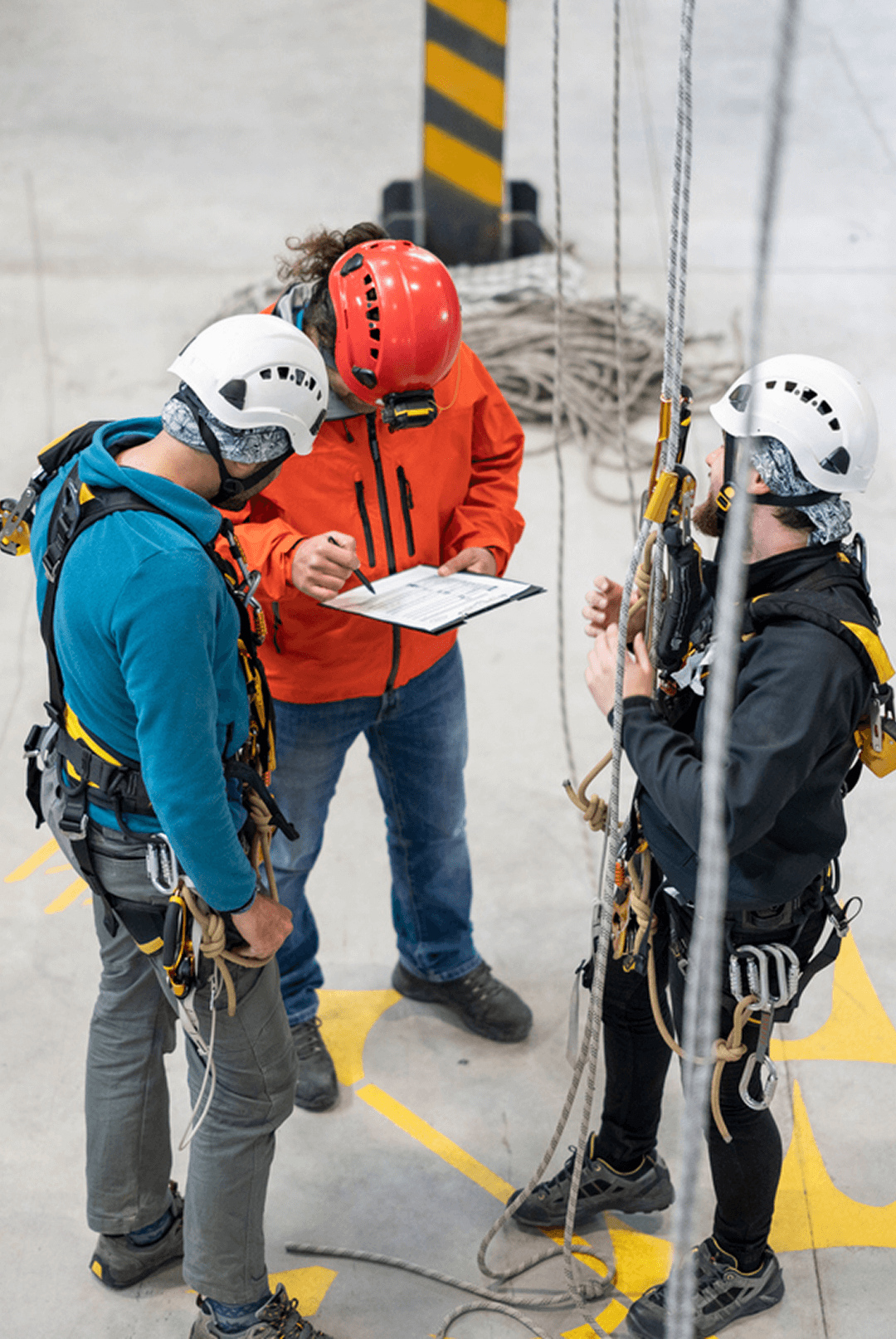 Drei Personen in Sicherheitsausrüstung besprechen Kletterausrüstung in einer Halle. Helme, Seile und Klemmbretter sichtbar.