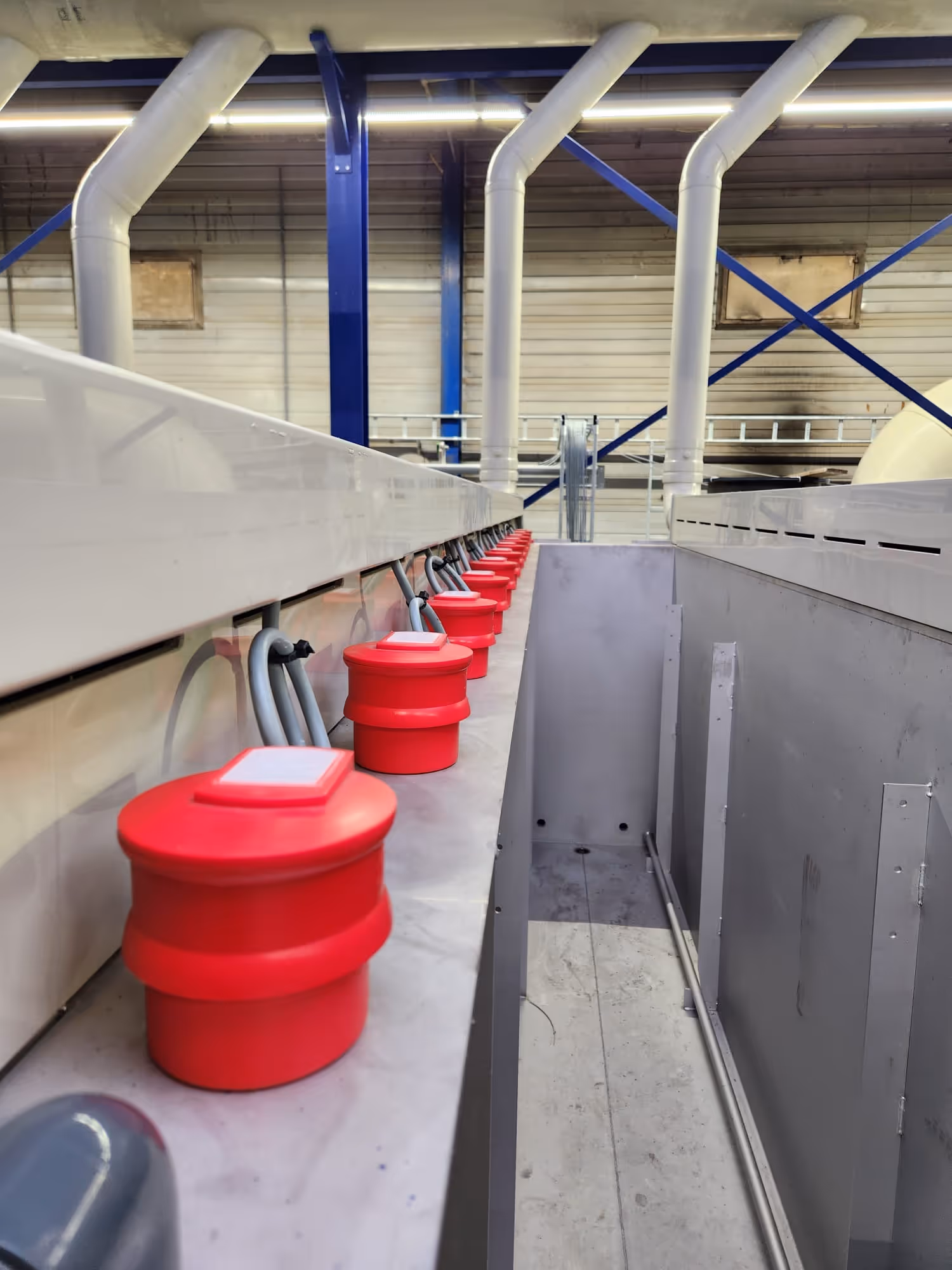 Rij rode schakelaars langs industriële apparatuur onder witte ventilatiebuizen in fabriekshal.