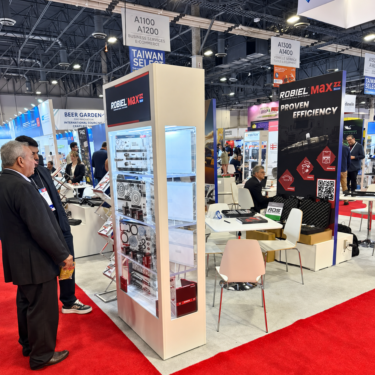 Trade show booth for Robiel Maxzz showcasing common rail system parts and promoting proven efficiency with product displays and informational banners, with attendees engaging nearby on a red carpeted floor.