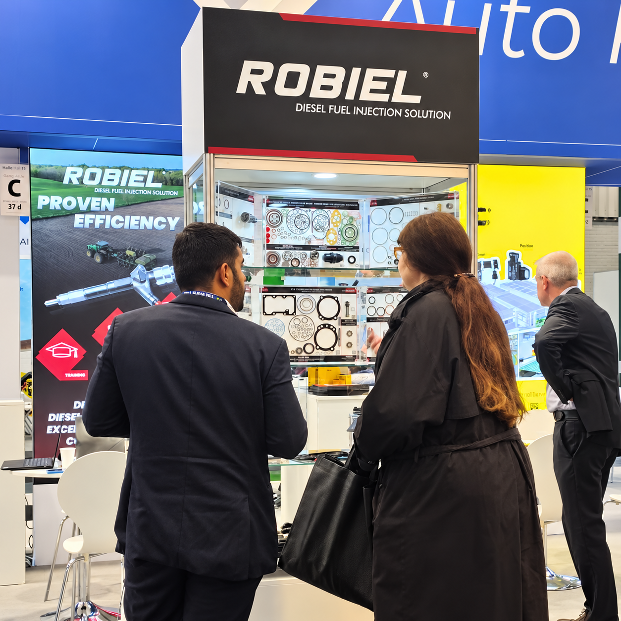 Three people in business attire stand at a trade show booth for Robiel, a diesel fuel injection solution company, examining display cases with mechanical parts and informational posters.