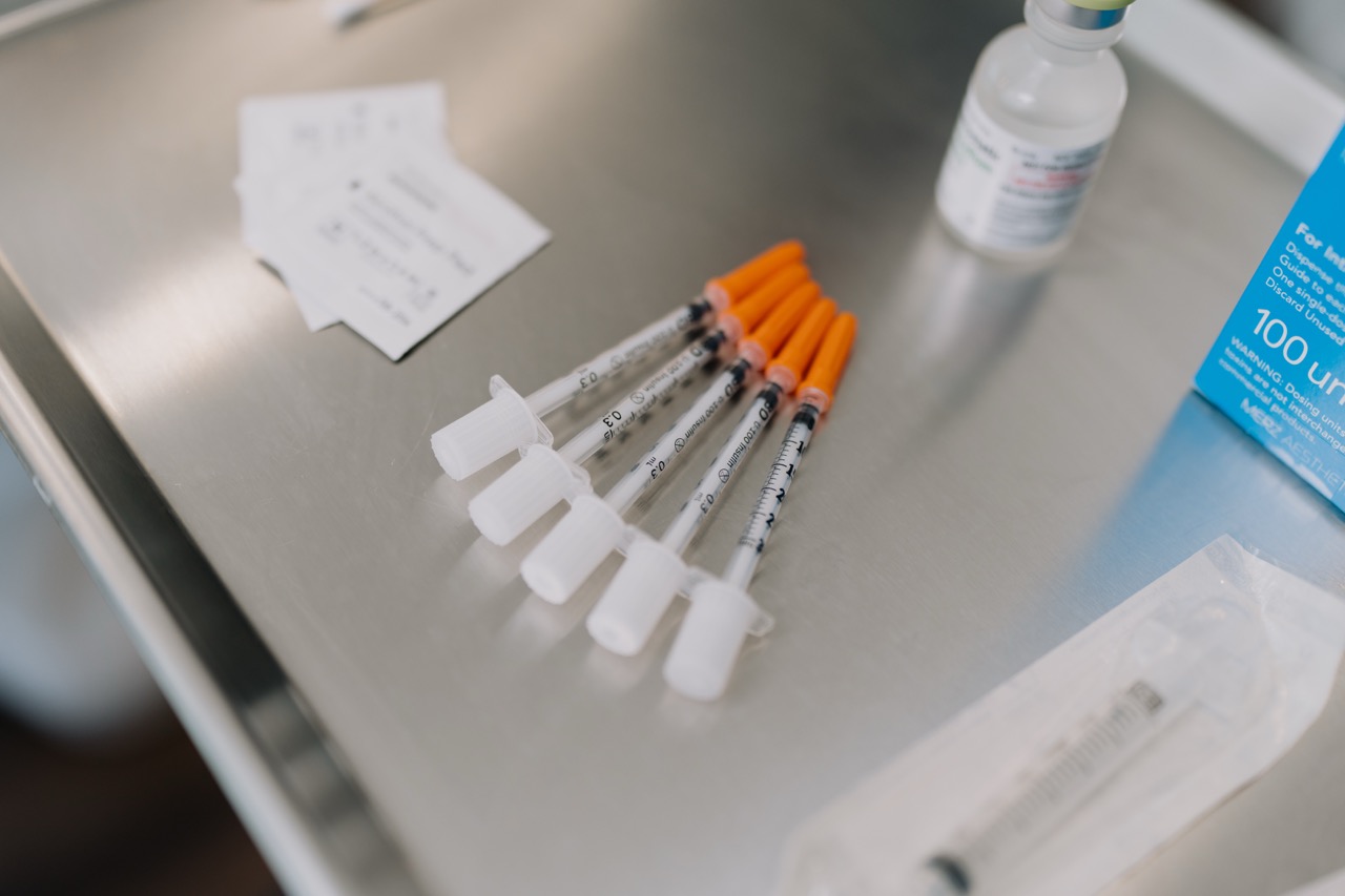 Six syringes with orange caps lined up on a metal medical tray with a vial and medication packaging nearby.