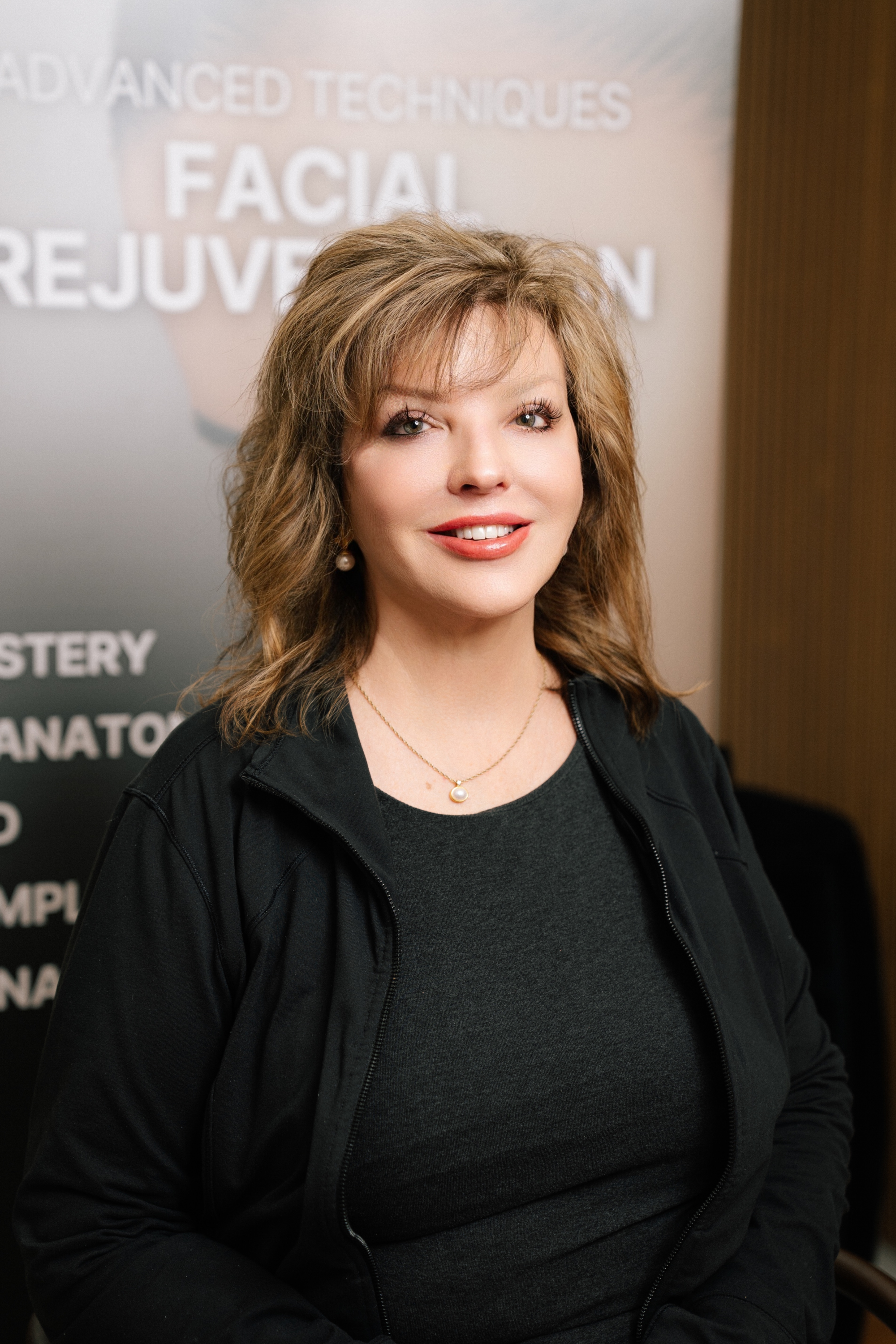 Middle-aged woman with shoulder-length blond hair wearing a black jacket and pearl necklace, smiling in front of a facial rejuvenation poster.