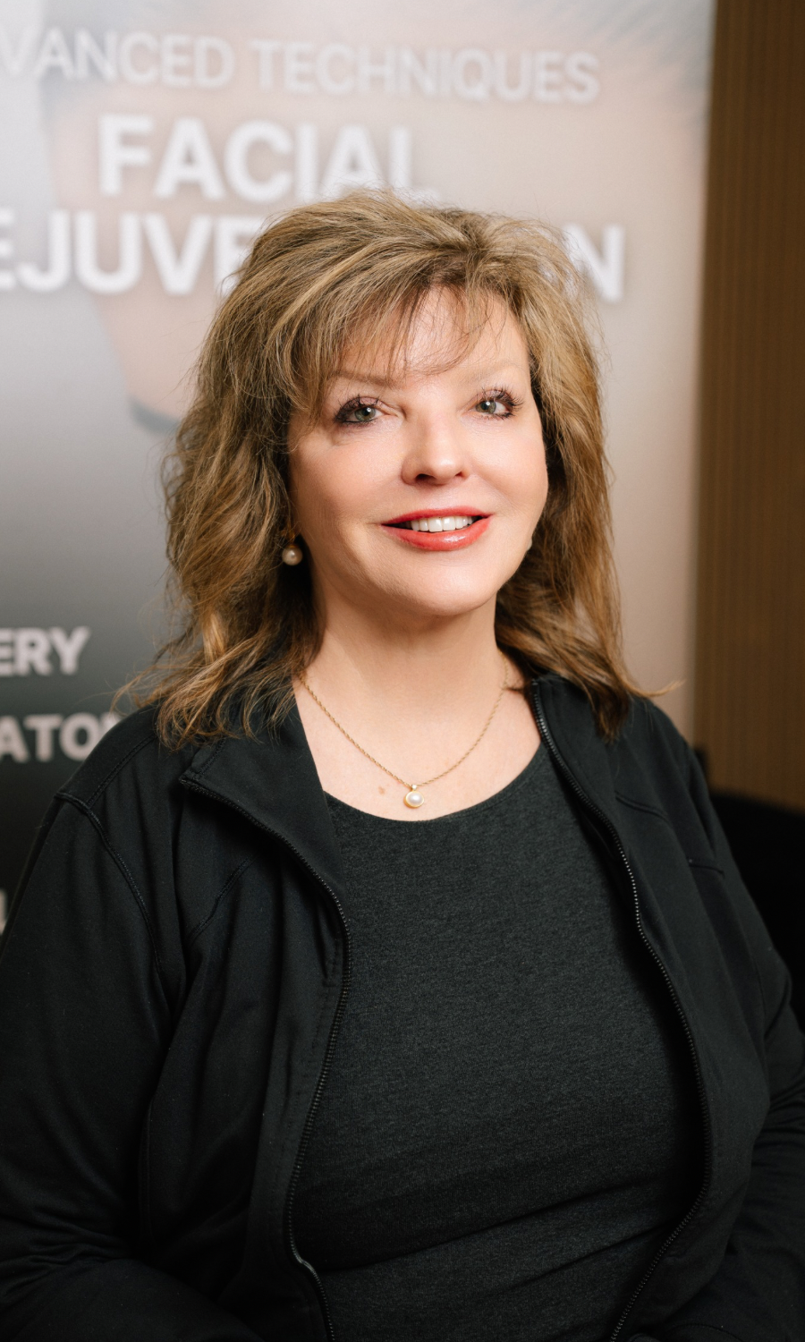 Middle-aged woman with shoulder-length blond hair wearing a black jacket and pearl necklace, smiling in front of a facial rejuvenation poster.