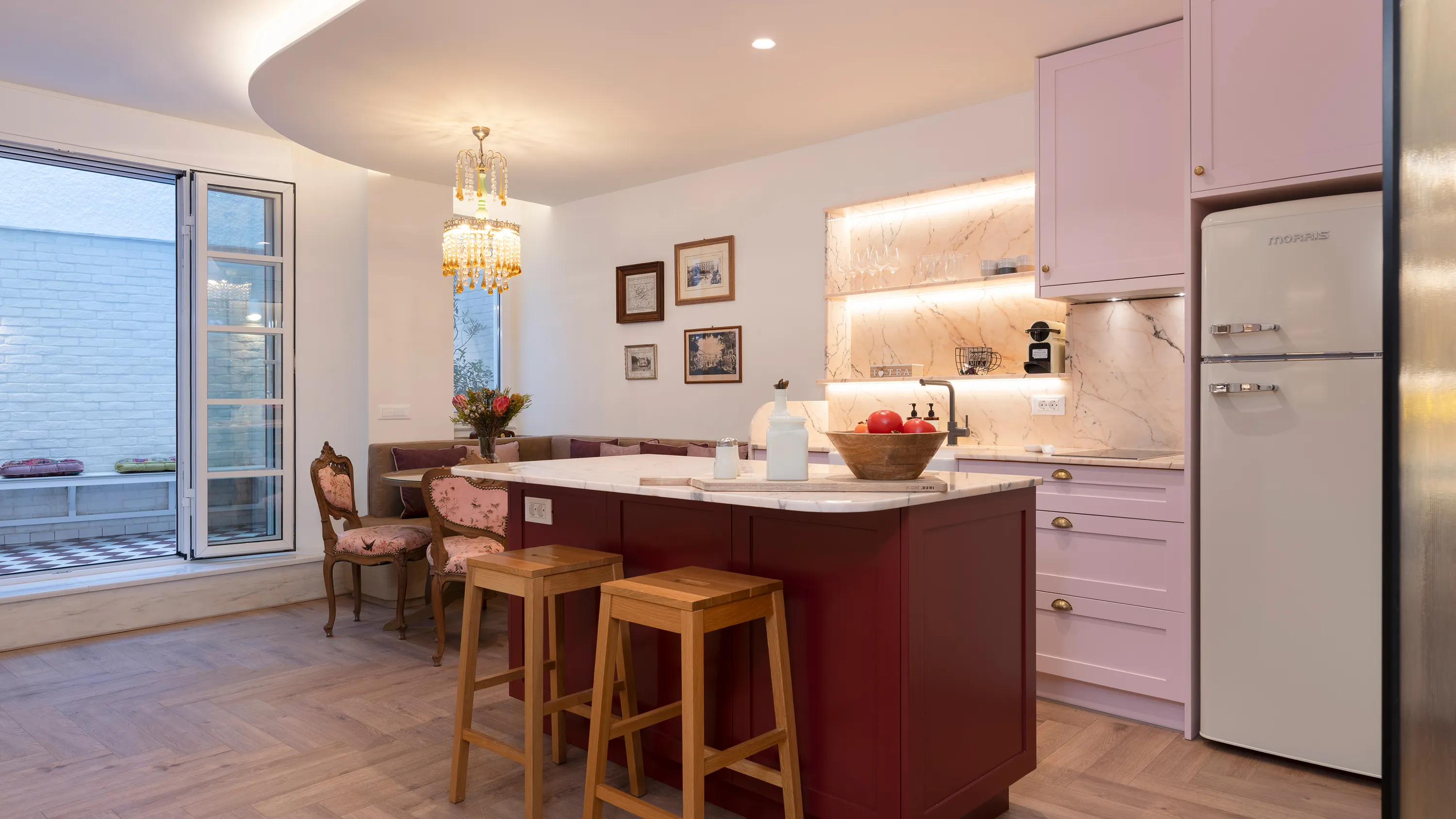 Modern kitchen with a burgundy island, two wooden stools, pink cabinets, vintage refrigerator, and a cozy seating area with floral chairs beneath a crystal chandelier.