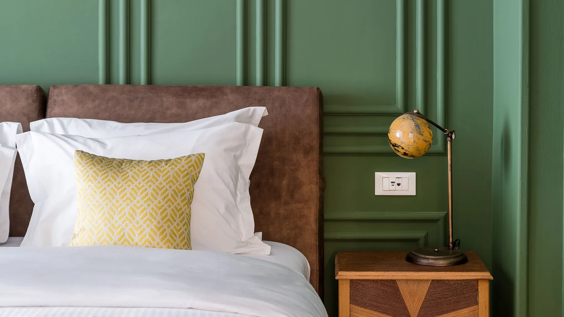 Close-up of a bed with white sheets, a yellow patterned pillow, brown headboard, green wall, wooden nightstand, and vintage-style yellow globe lamp.