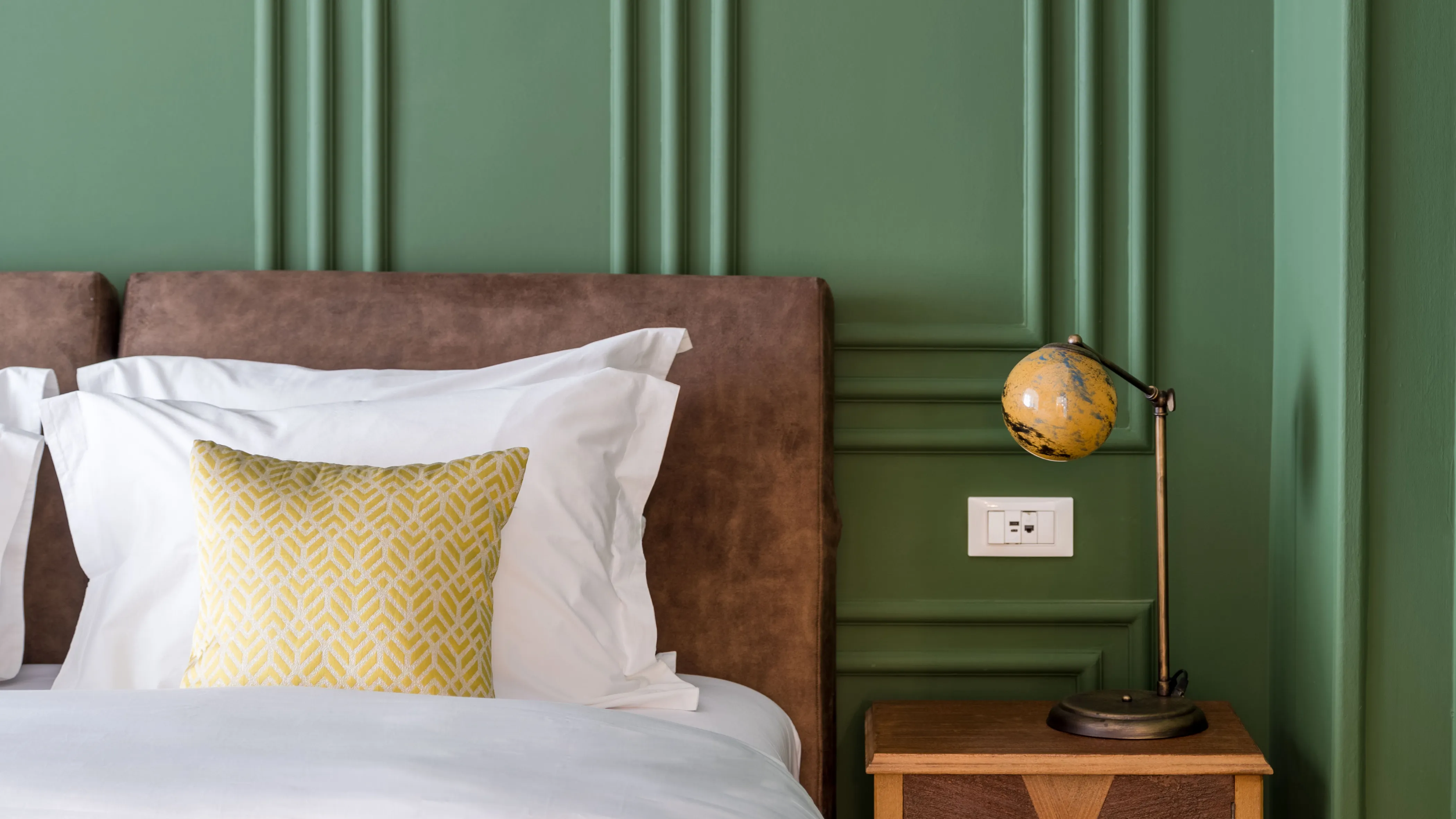 Close-up of a bed with white sheets, a yellow patterned pillow, brown headboard, green wall, wooden nightstand, and vintage-style yellow globe lamp.