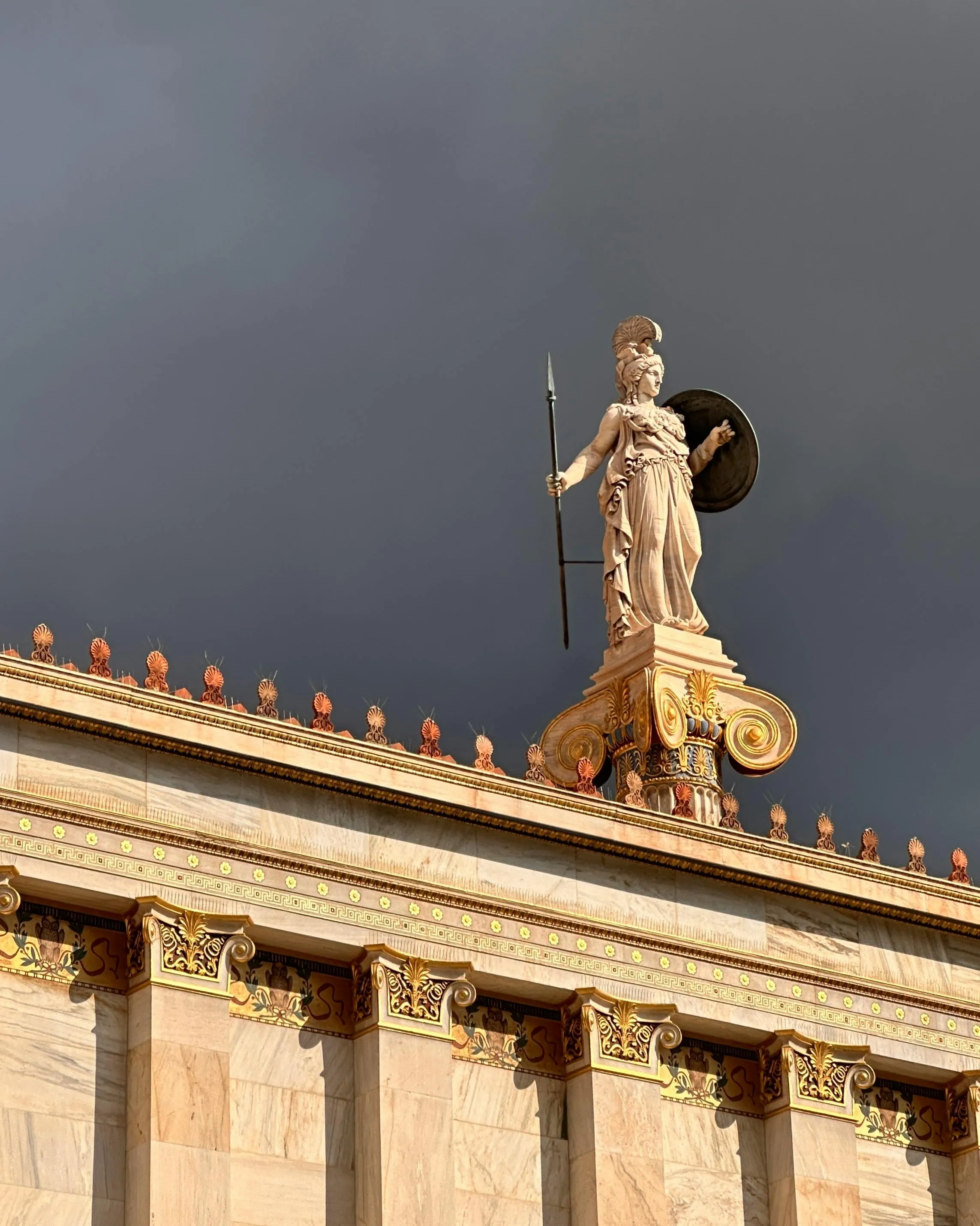 Statue of a warrior goddess holding a spear and shield atop a classical building with ornate columns against a dark cloudy sky.