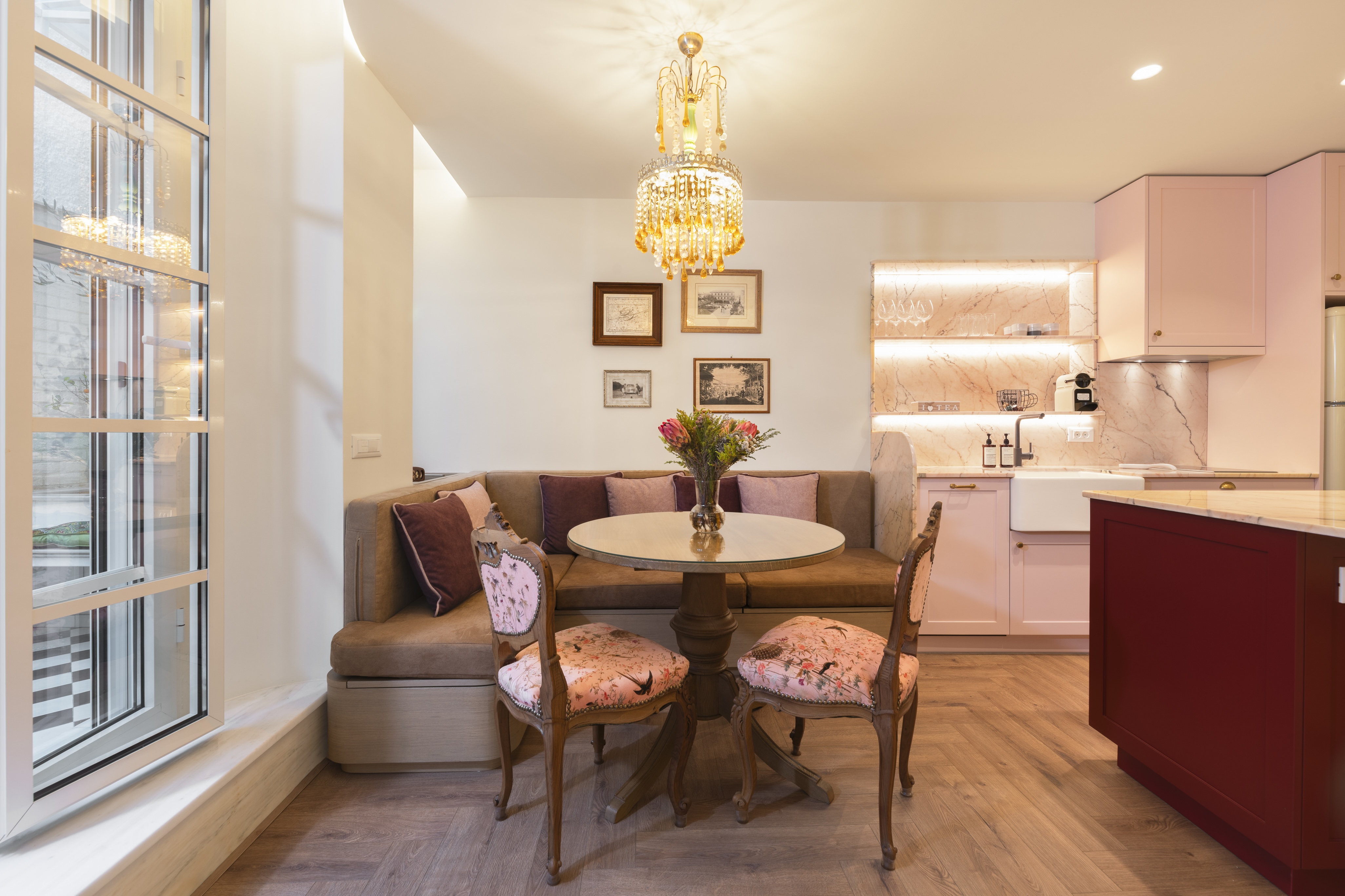 Modern kitchen with a burgundy island, two wooden stools, pink cabinets, vintage refrigerator, and a cozy seating area with floral chairs beneath a crystal chandelier.