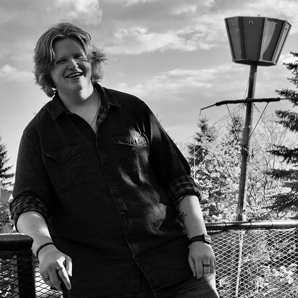 Smiling person with medium-length hair leaning on a railing outdoors with trees and sky in the background.
