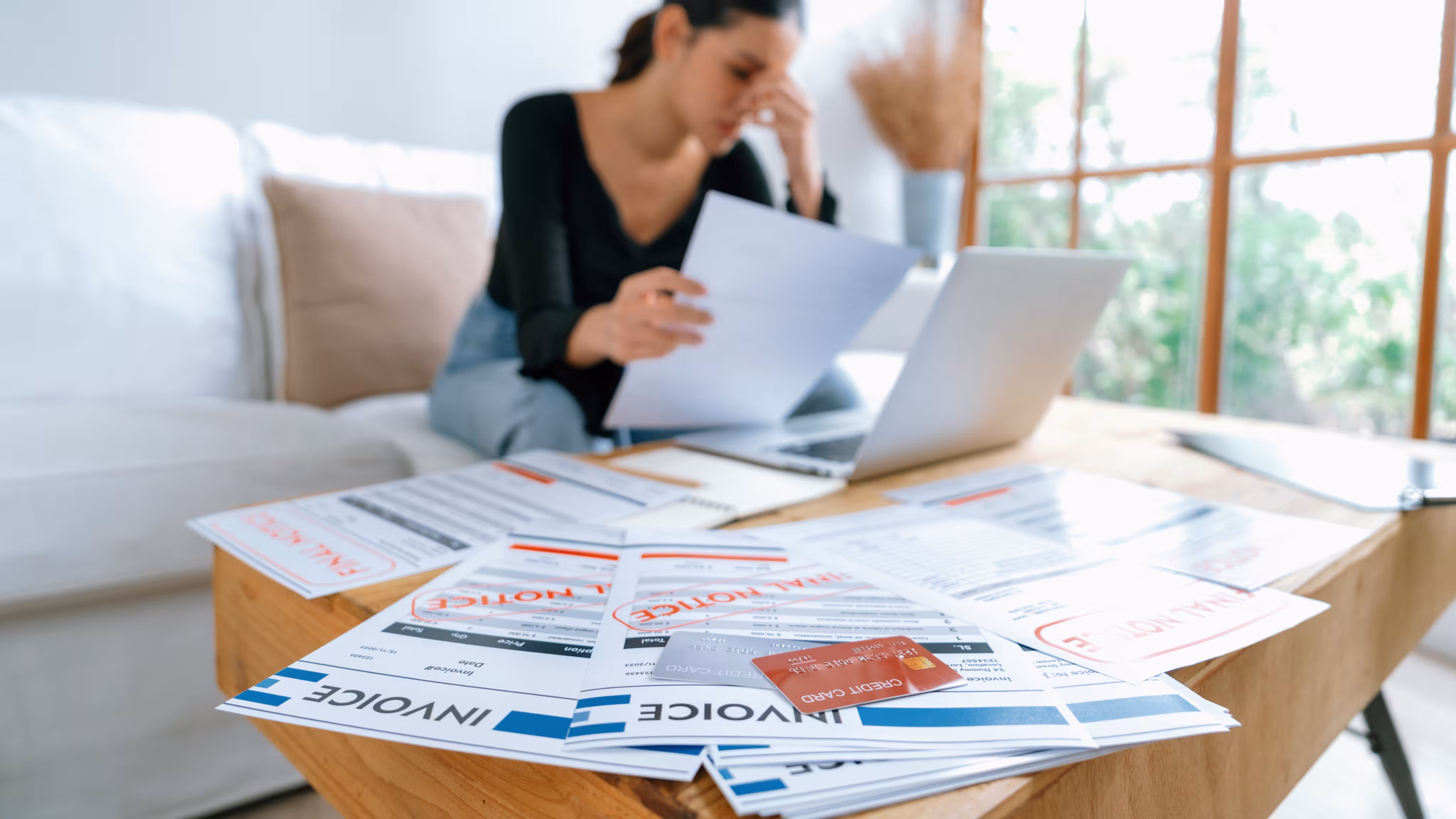 Woman sitting on a couch looking stressed while reviewing bills and invoices marked with 'FINAL NOTICE' on a table with a credit card and laptop.