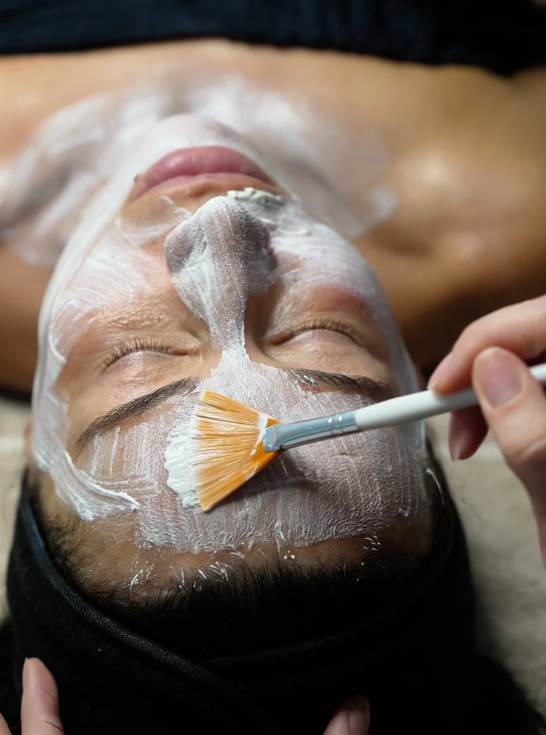 Person receiving a facial treatment with a white cream mask being applied on the forehead using a fan-shaped brush.
