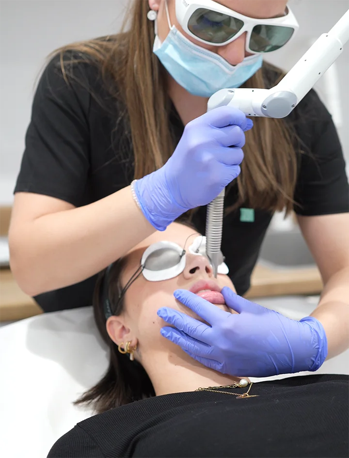 Une professionnelle en lunettes de protection et masque effectue un traitement au laser sur les lèvres d'une patiente allongée portant des lunettes de protection oculaires.