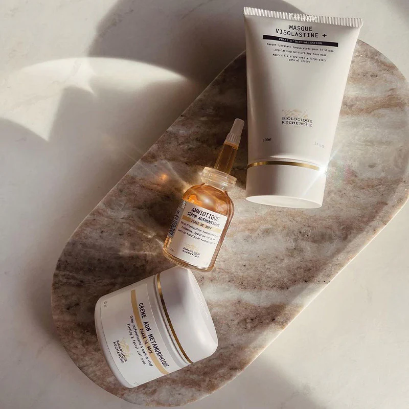 Trois produits de soins de la peau Biologique Recherche posés sur un plateau en pierre marron sous une lumière naturelle. clinique skinin