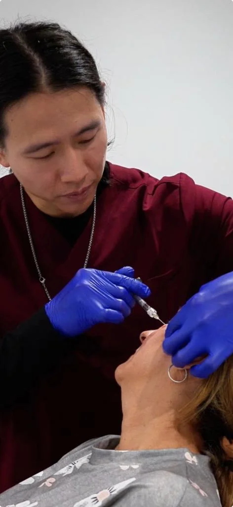 Un professionnel vêtu d'un uniforme rouge et portant des gants bleus injecte un produit dans le visage d'une patiente allongée.