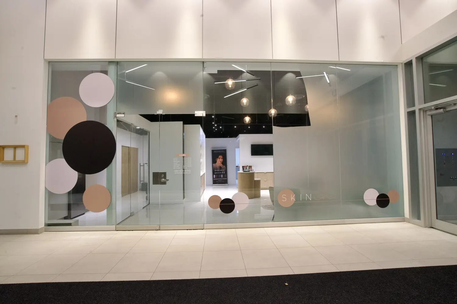 Façade vitrée de la clinique médico-esthétique SkinIn de la peau moderne avec des cercles décoratifs de différentes couleurs sur le verre et un intérieur lumineux avec comptoir et affiches. Brossard Quartier dix30