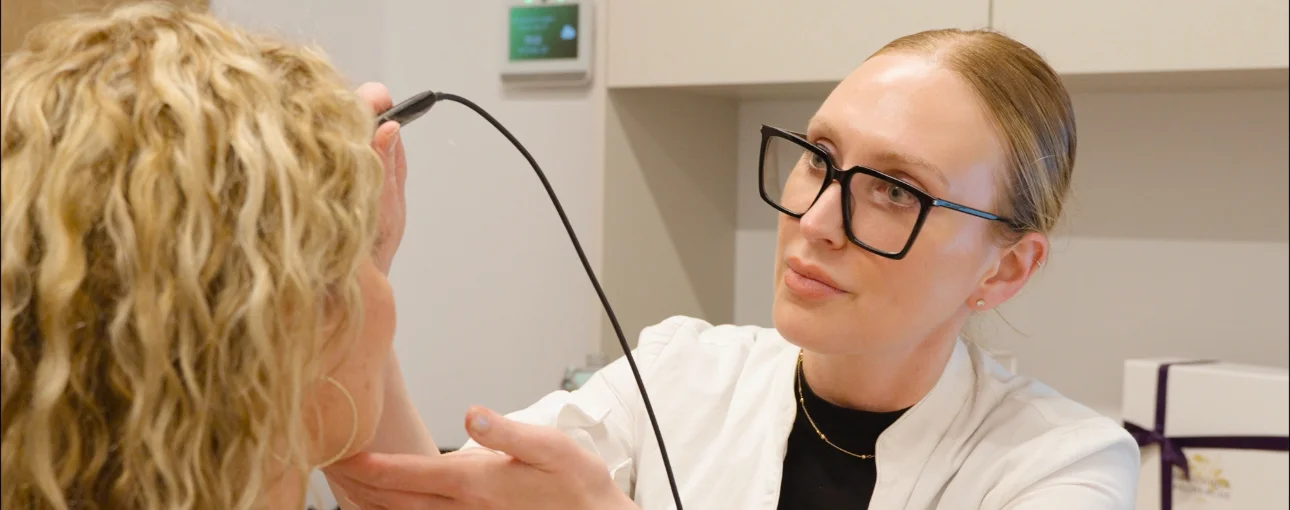 Une professionnelle en blouse blanche et lunettes noires utilise un appareil pour examiner la peau du visage d'une femme aux cheveux bouclés blonds.