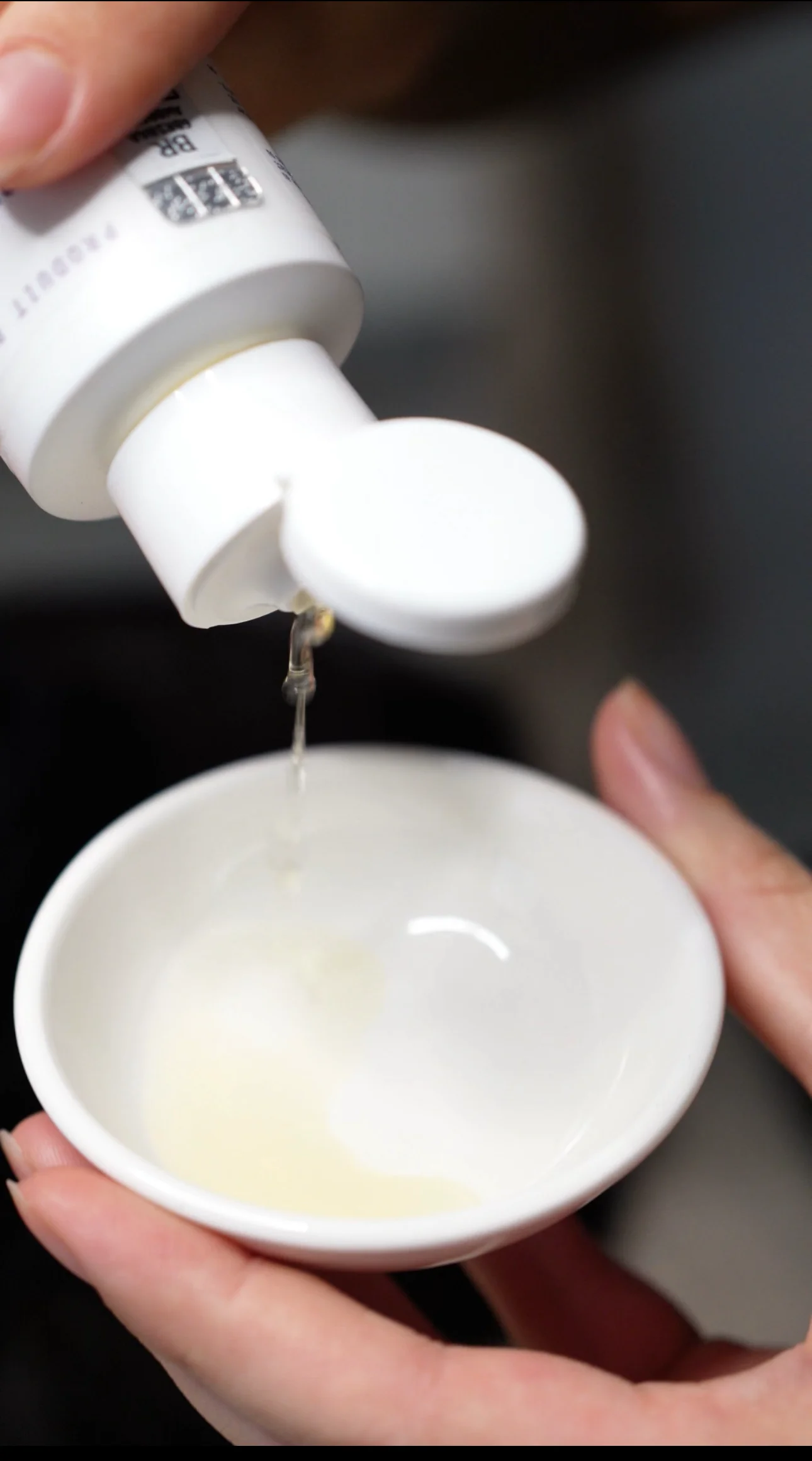 Liquide clair étant versé d'un flacon blanc dans un petit bol blanc tenu à la main.