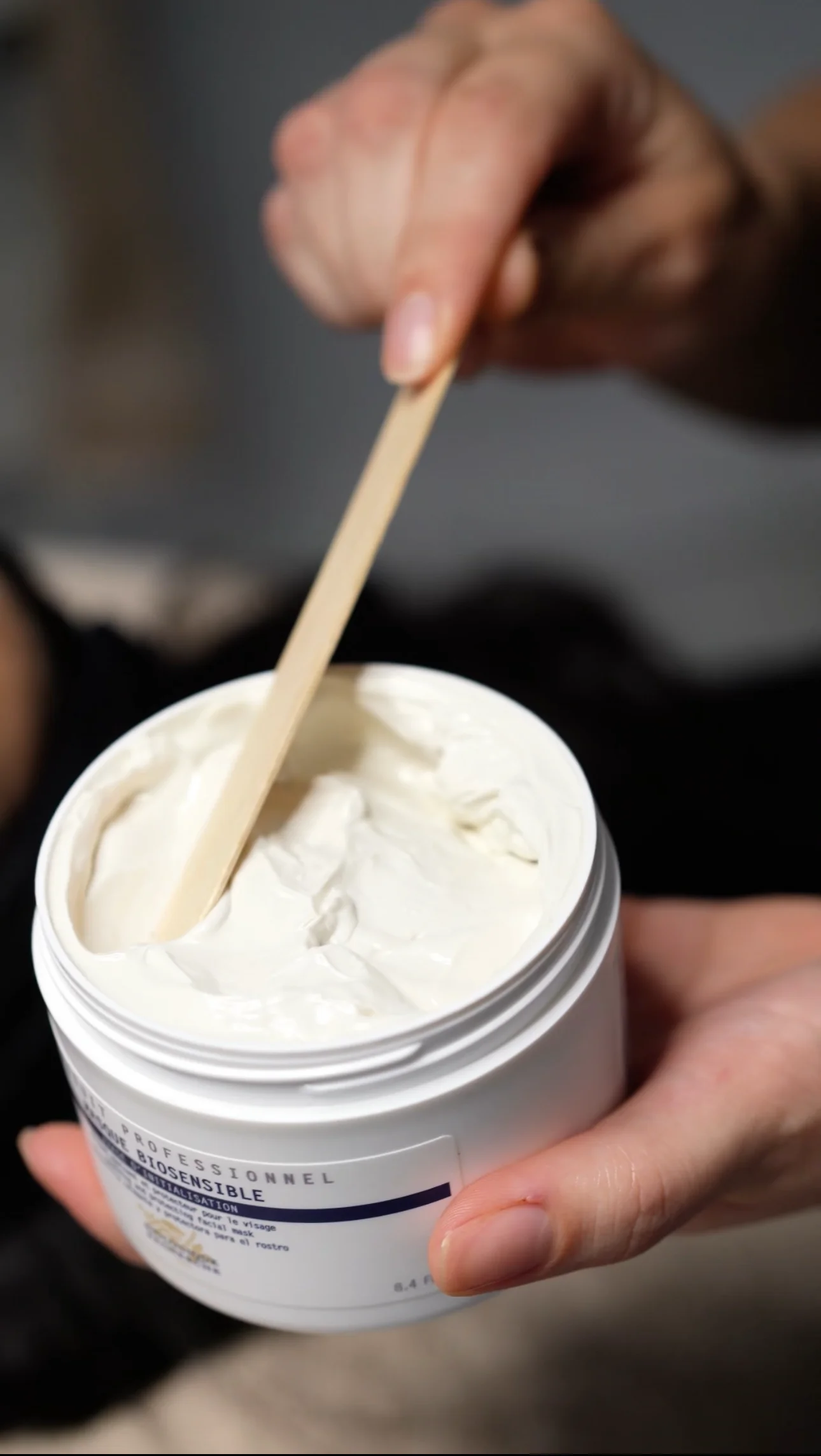 Main tenant un pot blanc de crème épaisse et un bâtonnet en bois inséré dans la crème.