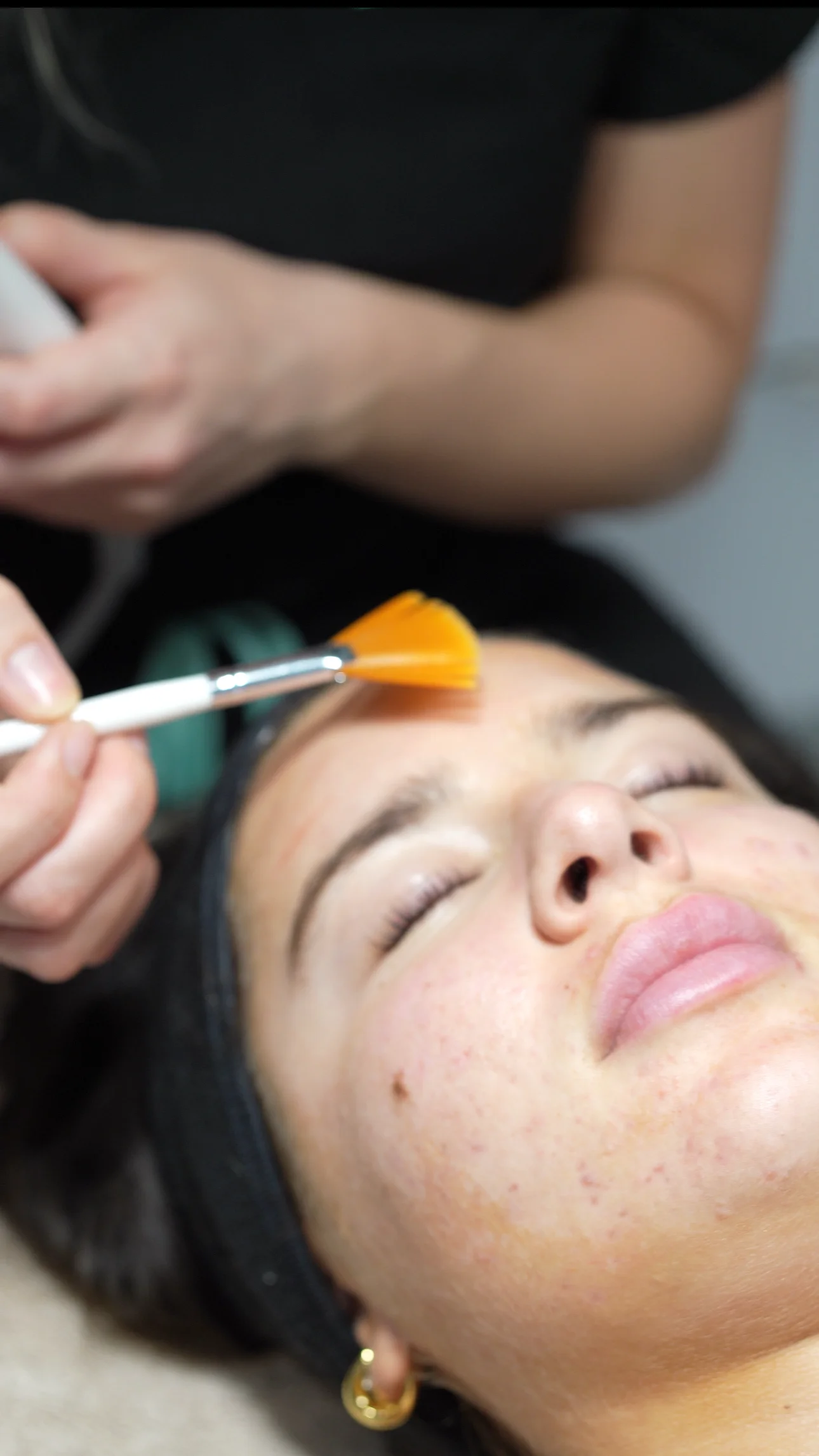 Une personne reçoit un soin du visage avec un pinceau appliqué sur le front, les yeux fermés et un bandeau noir tenant les cheveux.