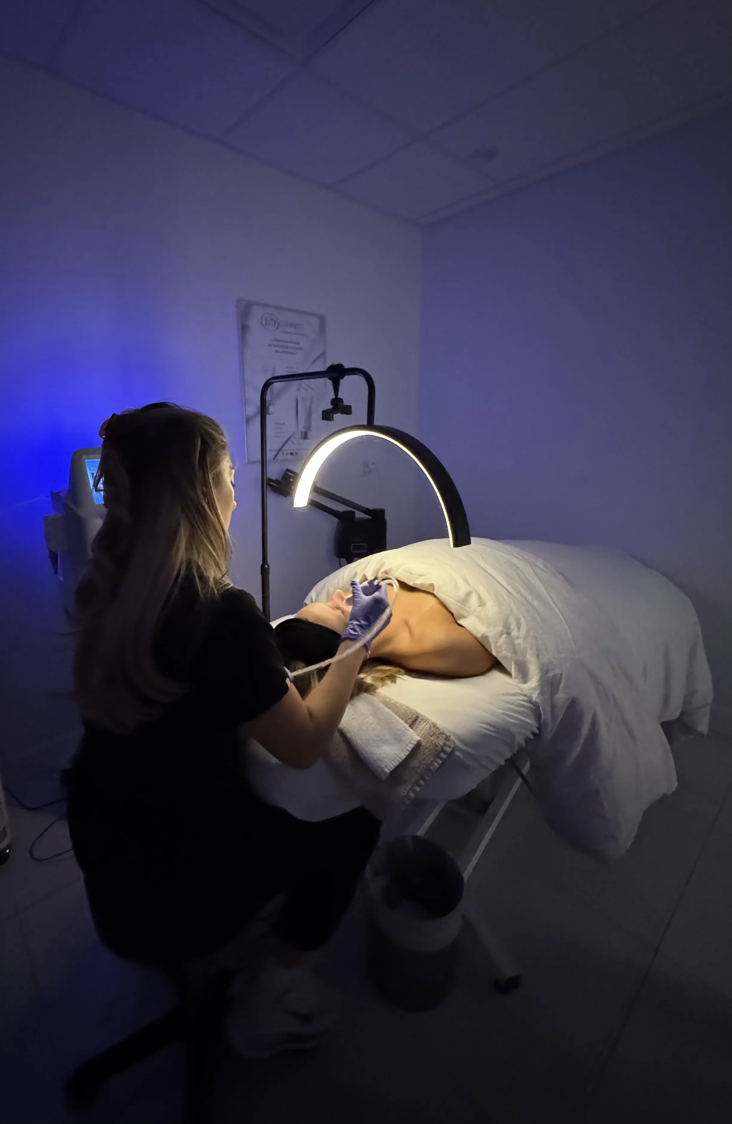 Esthéticienne réalisant un soin du visage à une cliente allongée sous une lumière annulaire dans une salle d'esthétique.