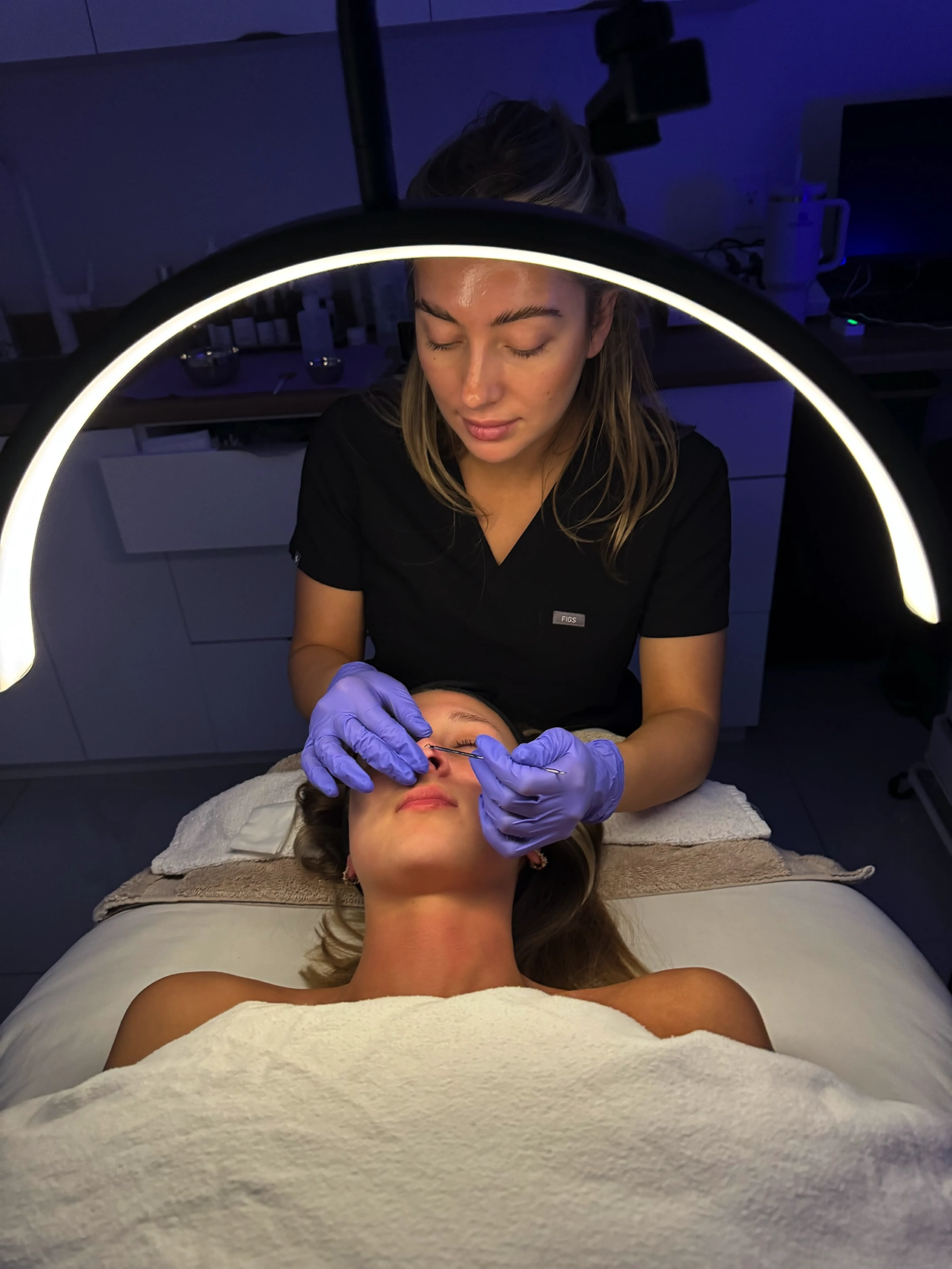 Esthéticienne portant des gants violets effectuant un soin du visage sous une lampe annulaire, une cliente allongée sous une couverture blanche. Charlie, clinique medico-esthétique rive-sud