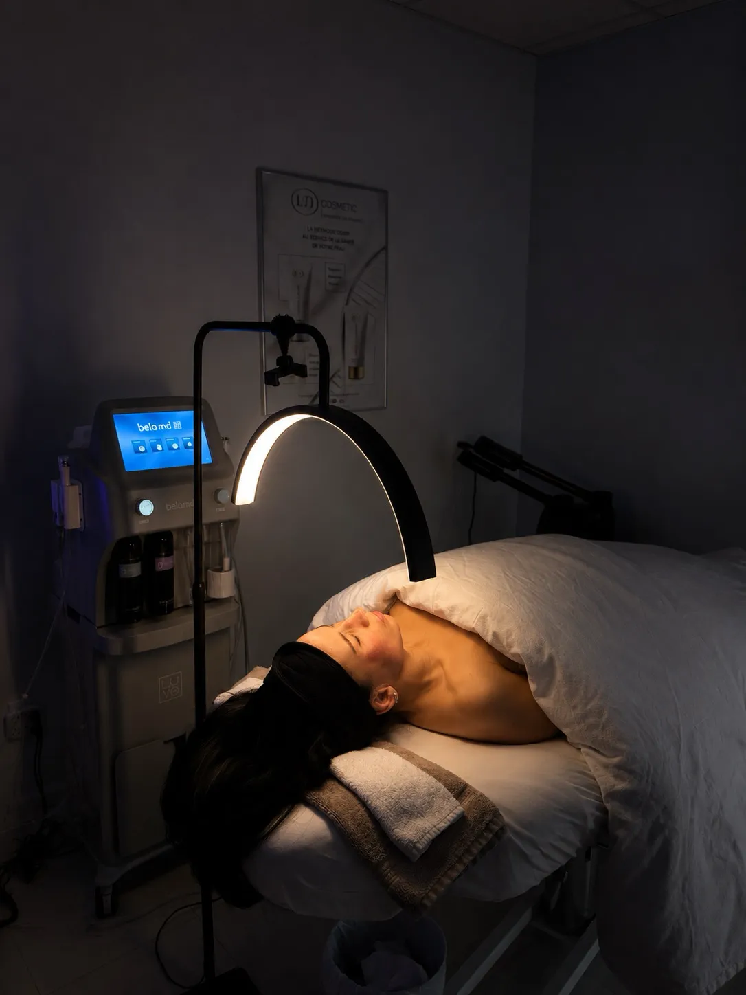 Femme allongée sous une couverture blanche recevant un soin du visage avec une lampe incurvée dans une cabine esthétique.
