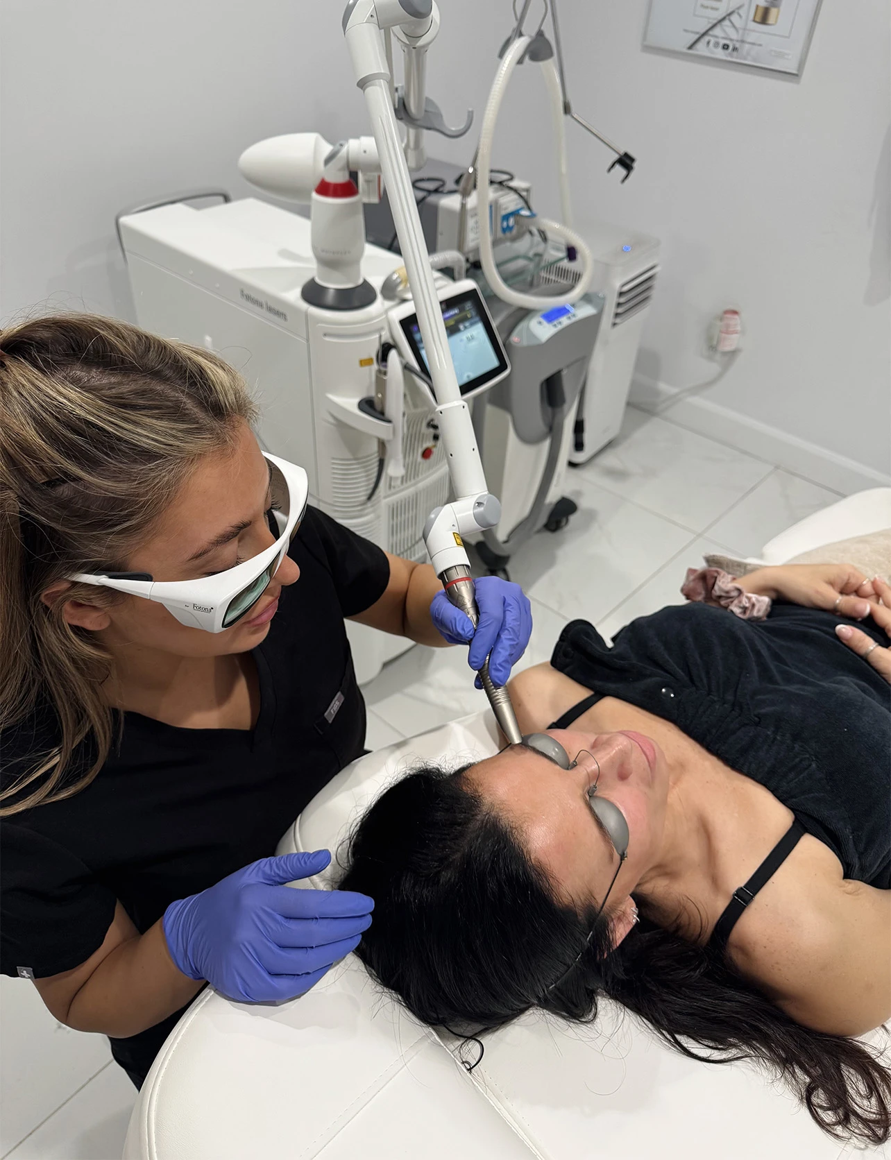 Professionnelle en lunettes de protection réalisant un traitement au laser sur le front d'une patiente allongée.
