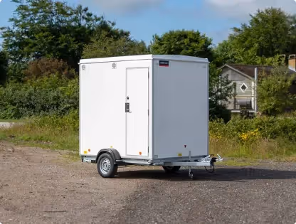 White rectangular referee house trailer with single door, parked on gravel near greenery and houses