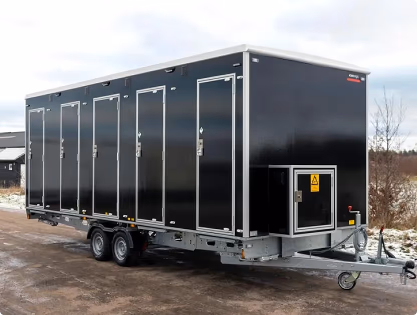 Black custom shower trailer with six individual access doors and a temperature control unit on front panel, parked outdoors on a dirt surface