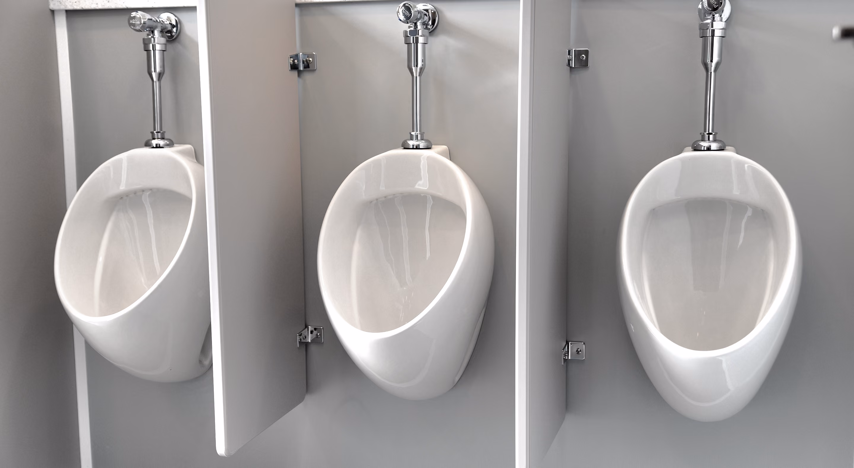 Three white urinals mounted on a gray wall with metal flush valves and privacy partitions.