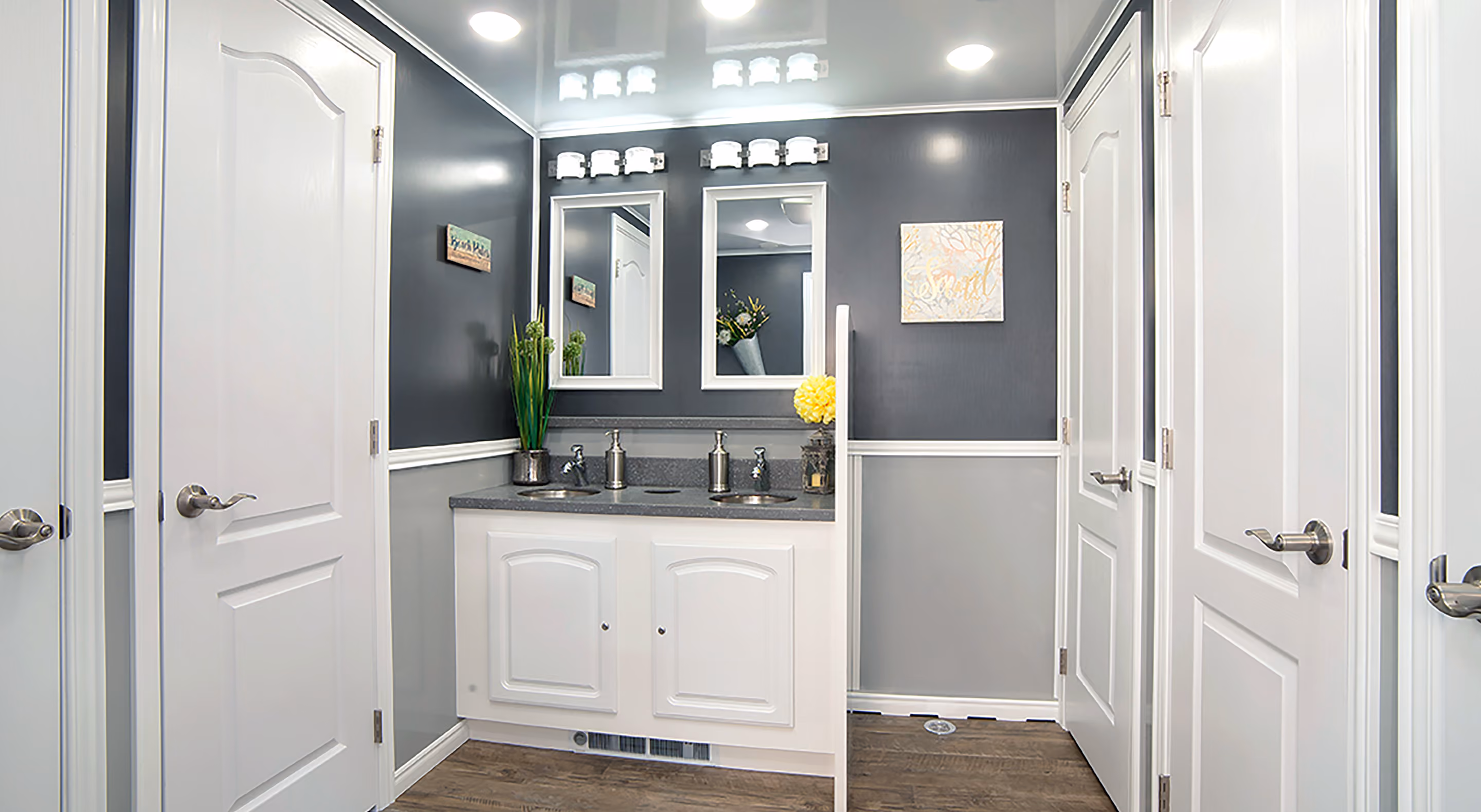Modern bathroom with dual sinks, decorative mirrors, and grey walls. White doors and wooden floor.