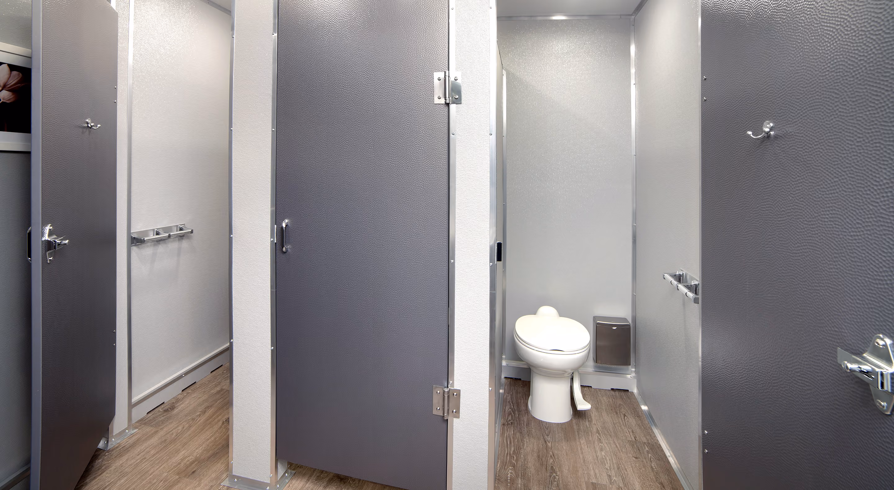Interior of a clean public restroom with gray stalls and wood-style flooring, one open showing a toilet.
